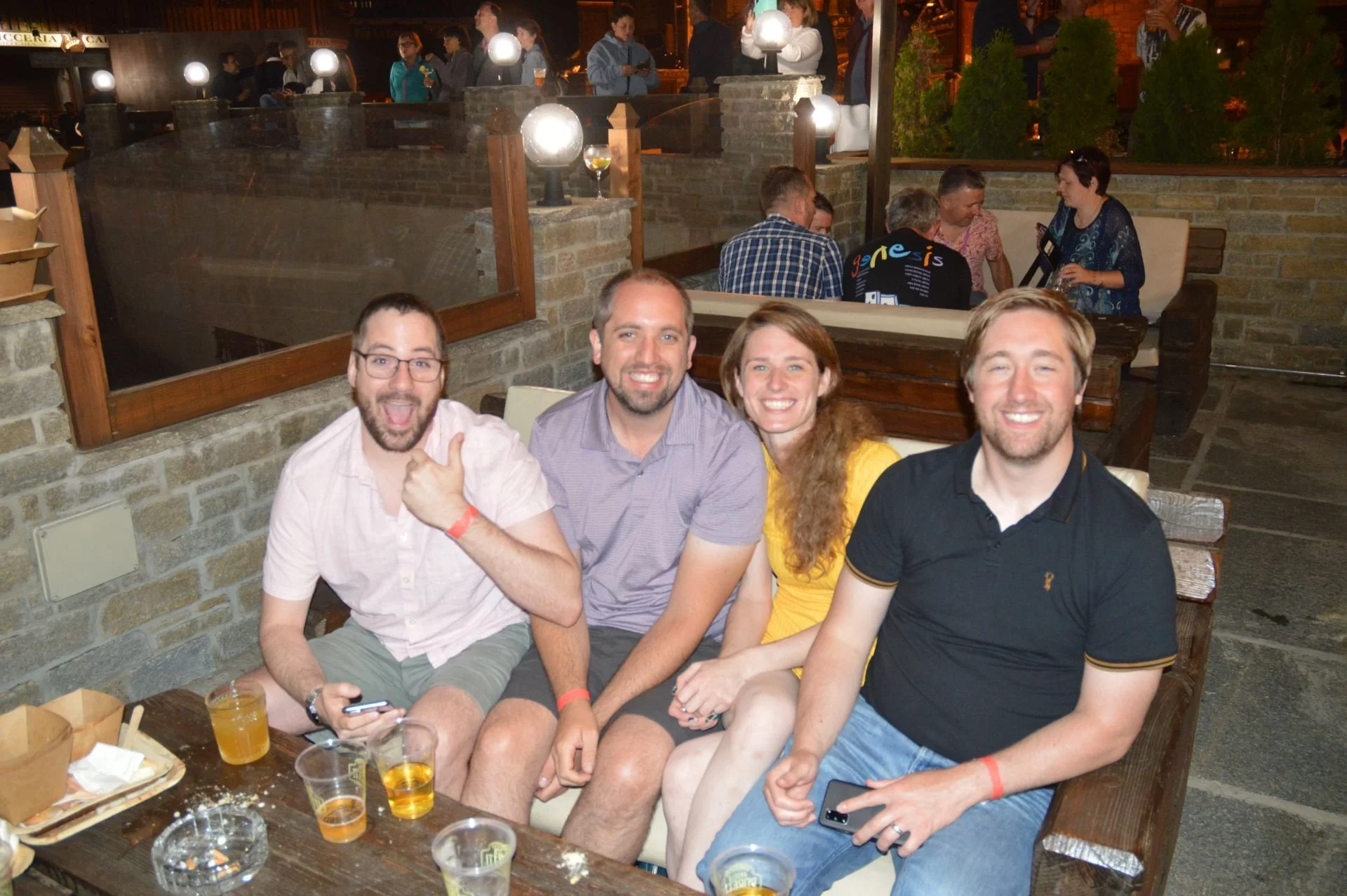 Group of four friends smiling and sitting on a bench at an outdoor restaurant during an evening gathering, with drinks on the table.