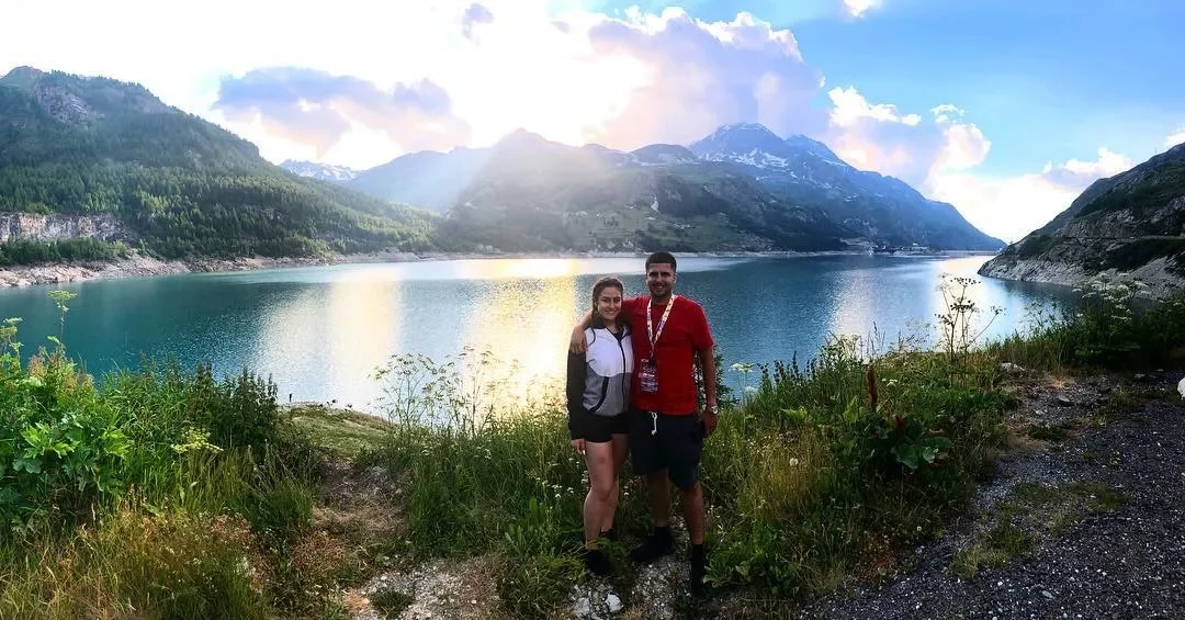 A man and woman standing together near a lake with mountains in the background, greenery, and a partly cloudy sky.