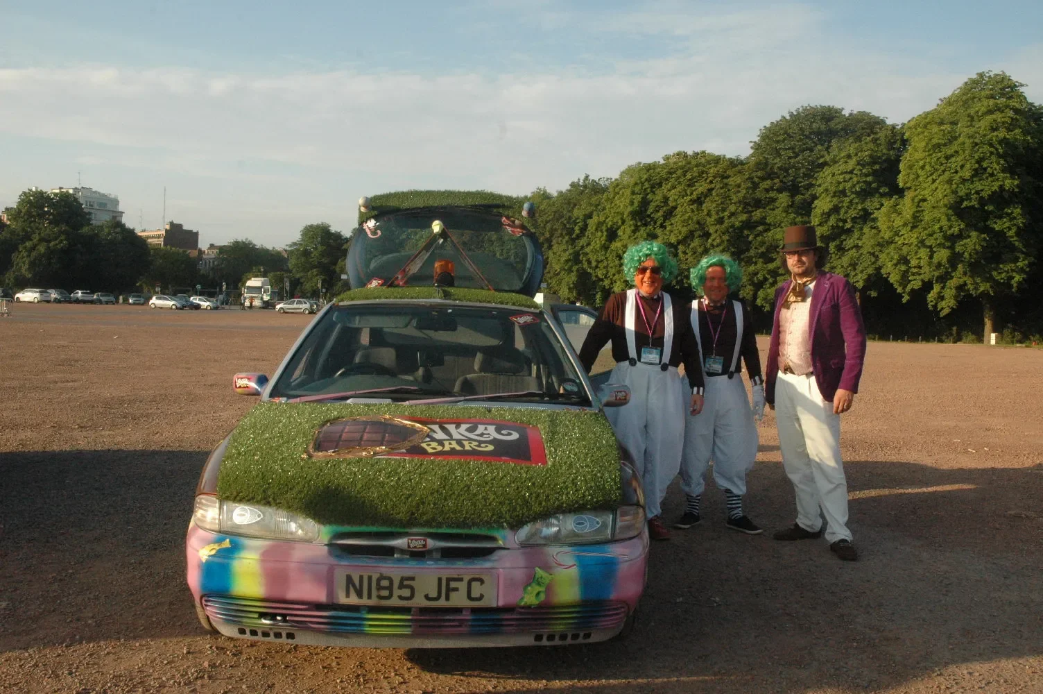 Three people dressed in clown costumes with bright green wigs, white pants, and suspenders standing next to a decorated car covered in grass-like material and colorful spray paint, in an open outdoor area with trees and buildings in the background.