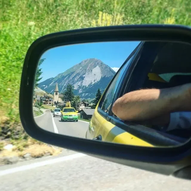 Scenic mountain view seen through a car side mirror, with a small village and cars on the road.