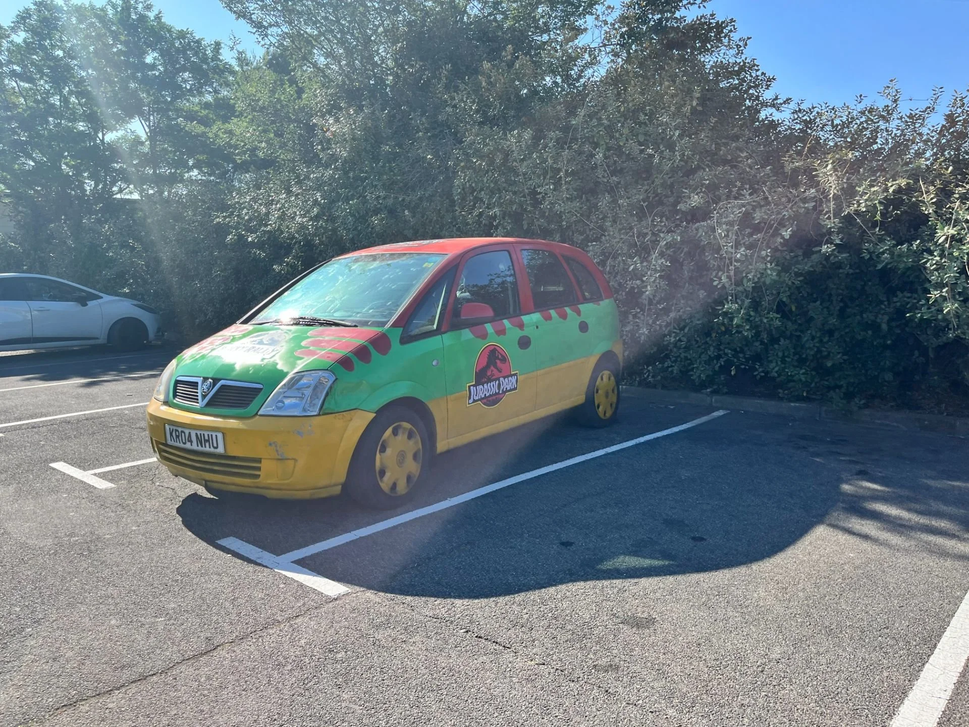 A small car decorated with a Jurassic Park dinosaur theme parked in a parking lot, with a white car nearby and trees in the background.
