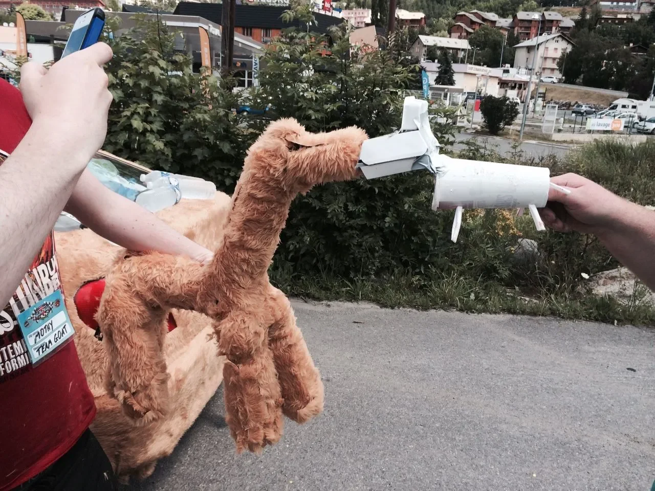 Someone holding a homemade, rocket-shaped craft aimed at a plush giraffe held by a person. The scene is outdoors with bushes, buildings, and parked cars in the background.
