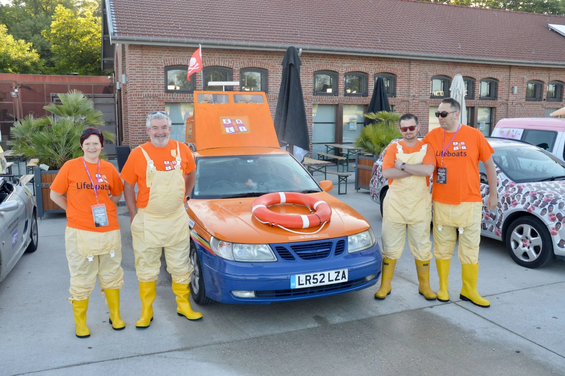Five people dressed as lifeguards standing in front of a small rescue boat on a blue and orange car, all wearing yellow boots, with a brick building and parked cars in the background.
