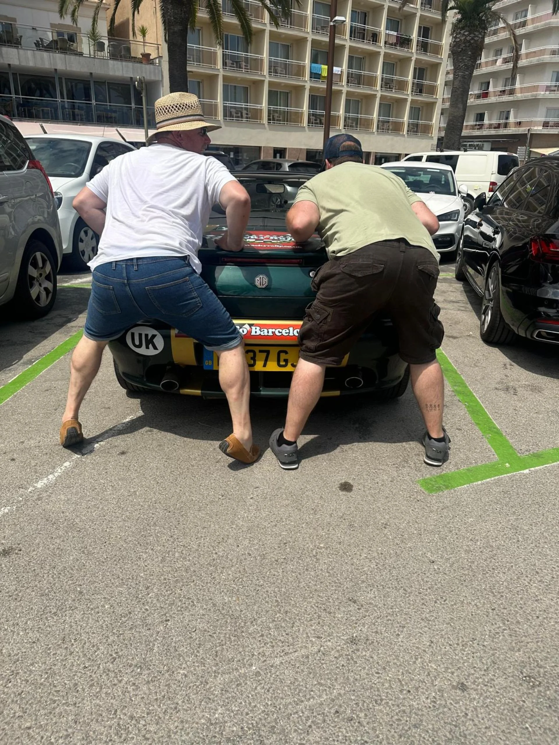 Two men leaning into the trunk of a black convertible car in a parking lot under sunny weather with palm trees and apartment buildings in the background.