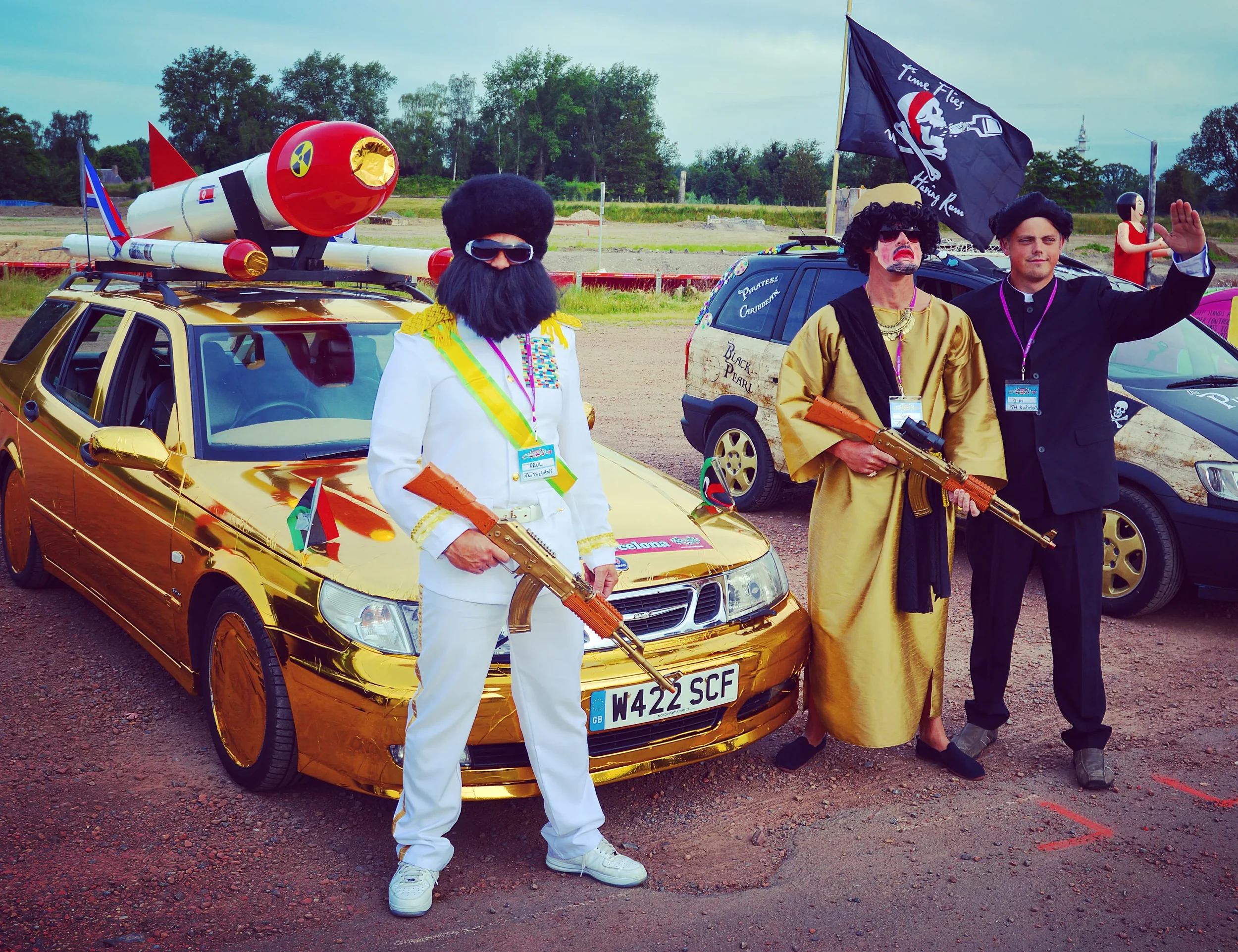 Three individuals dressed as famous figures in front of a gold car, holding toy guns, during a themed event or parade. One person is dressed as a military leader with a white uniform and medals, another as a rapper in a gold robe with a toy gun, and 