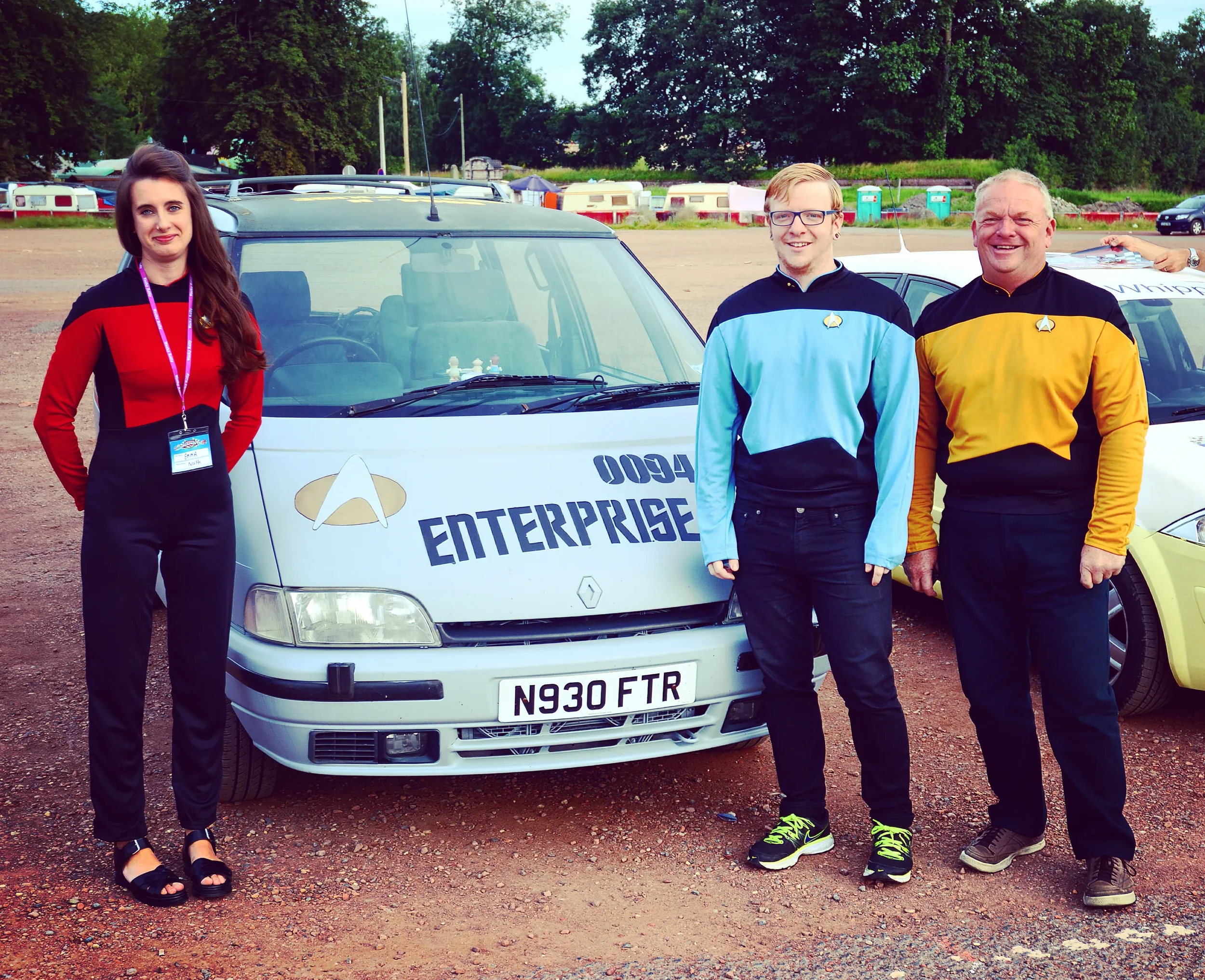 Group of three people standing in front of a white Enterprise car, two men in Star Trek uniforms and a woman with a badge, outdoors on a cloudy day