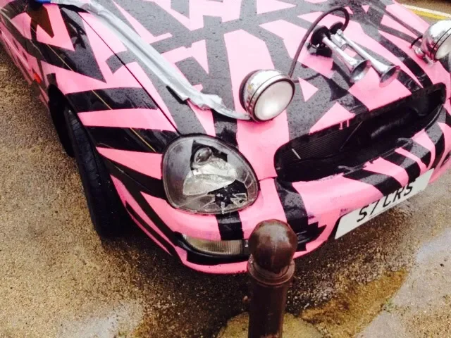 Pink and black sports car with racing stripes, front headlight, fog lights, and a license plate reading 'S1CK3' parked near a metal post on a wet pavement.