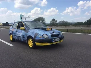 A blue vintage-style car with yellow wheels on a highway, with sky and clouds in the background.
