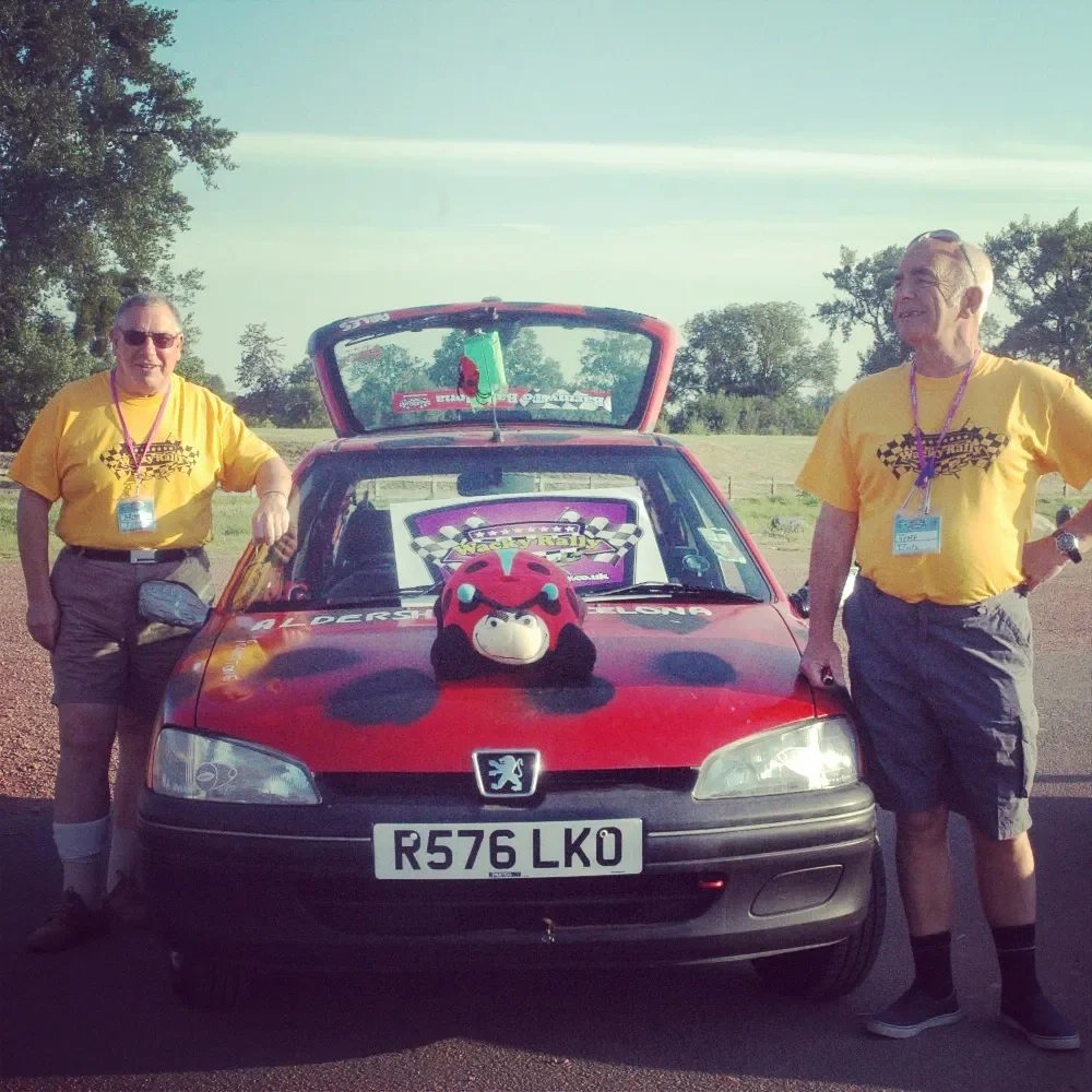 Two men in yellow t-shirts standing beside a red car with a plush toy on the hood, open trunk, and a racing flag sticker. The scene appears to be at a racing event or gathering, with trees and open space in the background.