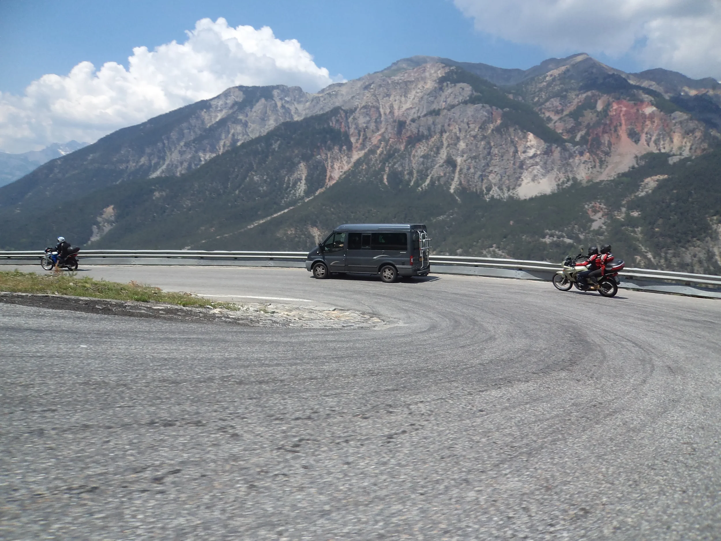 Motorcycle and van on a mountain road with a mountain range in the background.