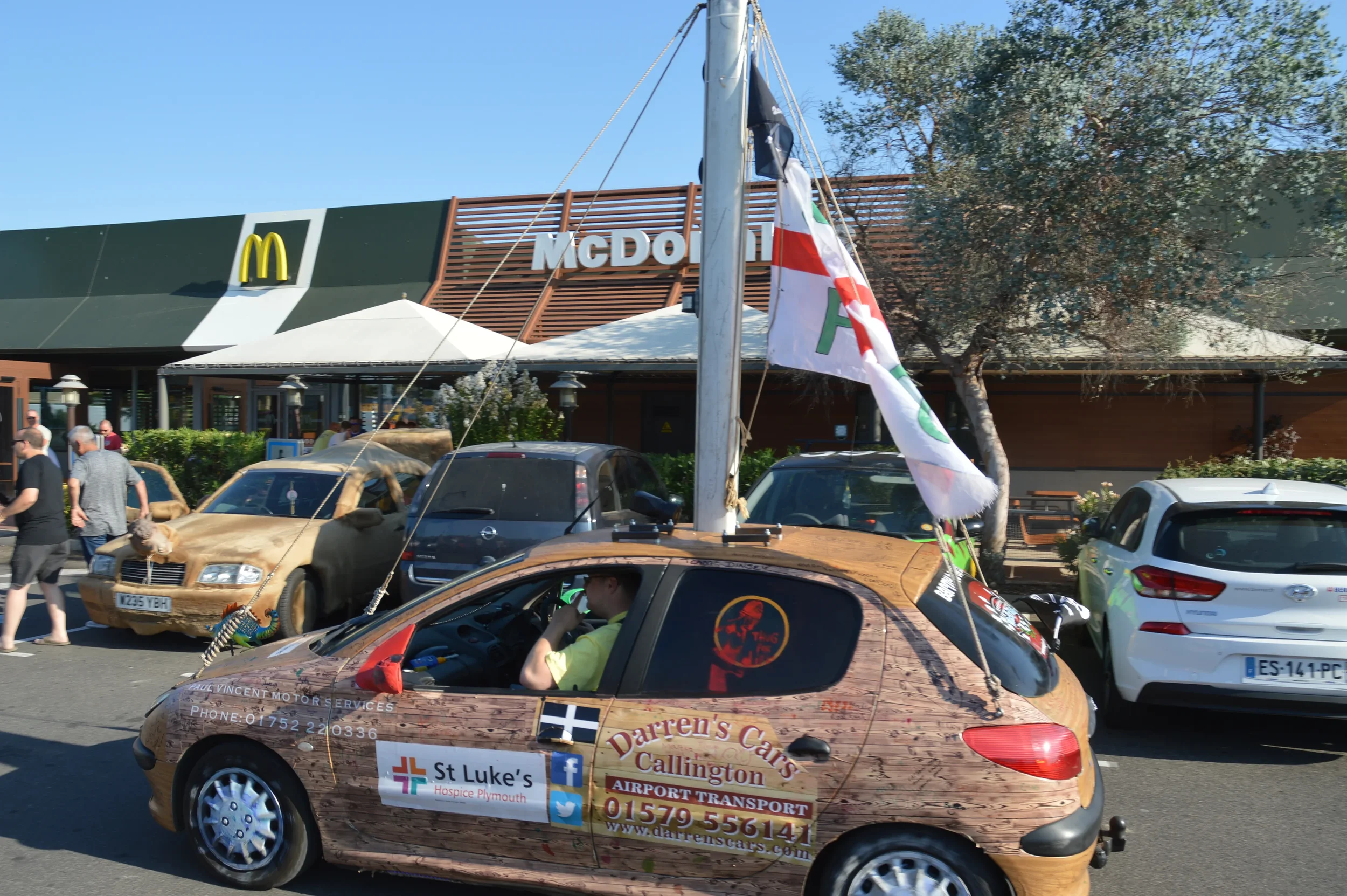 A small car decorated to look like wood with advertisements and signs for Darren's Cars Callington on the side, and a flag on the roof. It is parked in front of a McDonald's restaurant, with other cars and people walking nearby, and a McDonald's sign