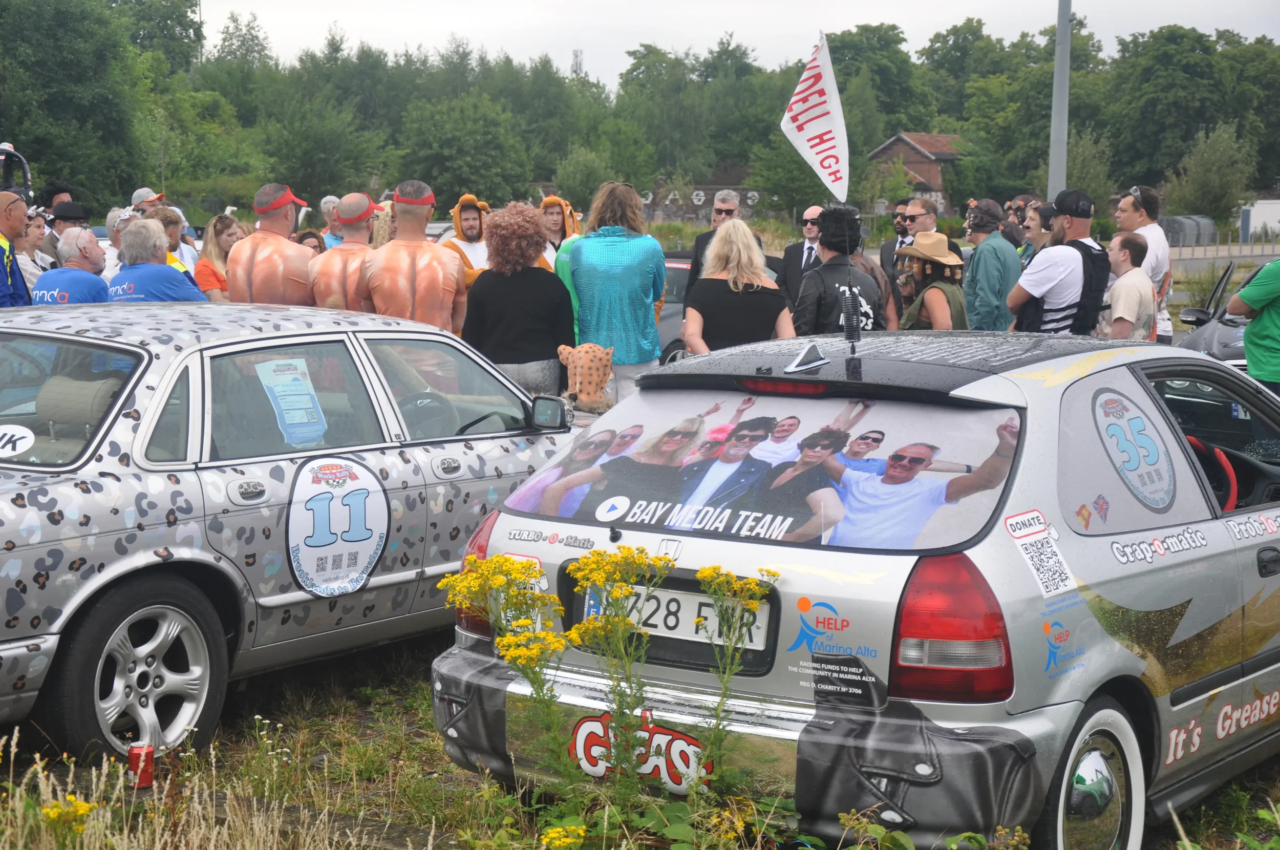 A crowd gathered around two cars, one decorated with leopard print and the other with popular club stickers. The cars are parked outdoors in a grassy area with yellow flowers, surrounded by trees and some buildings in the background. Many people are 