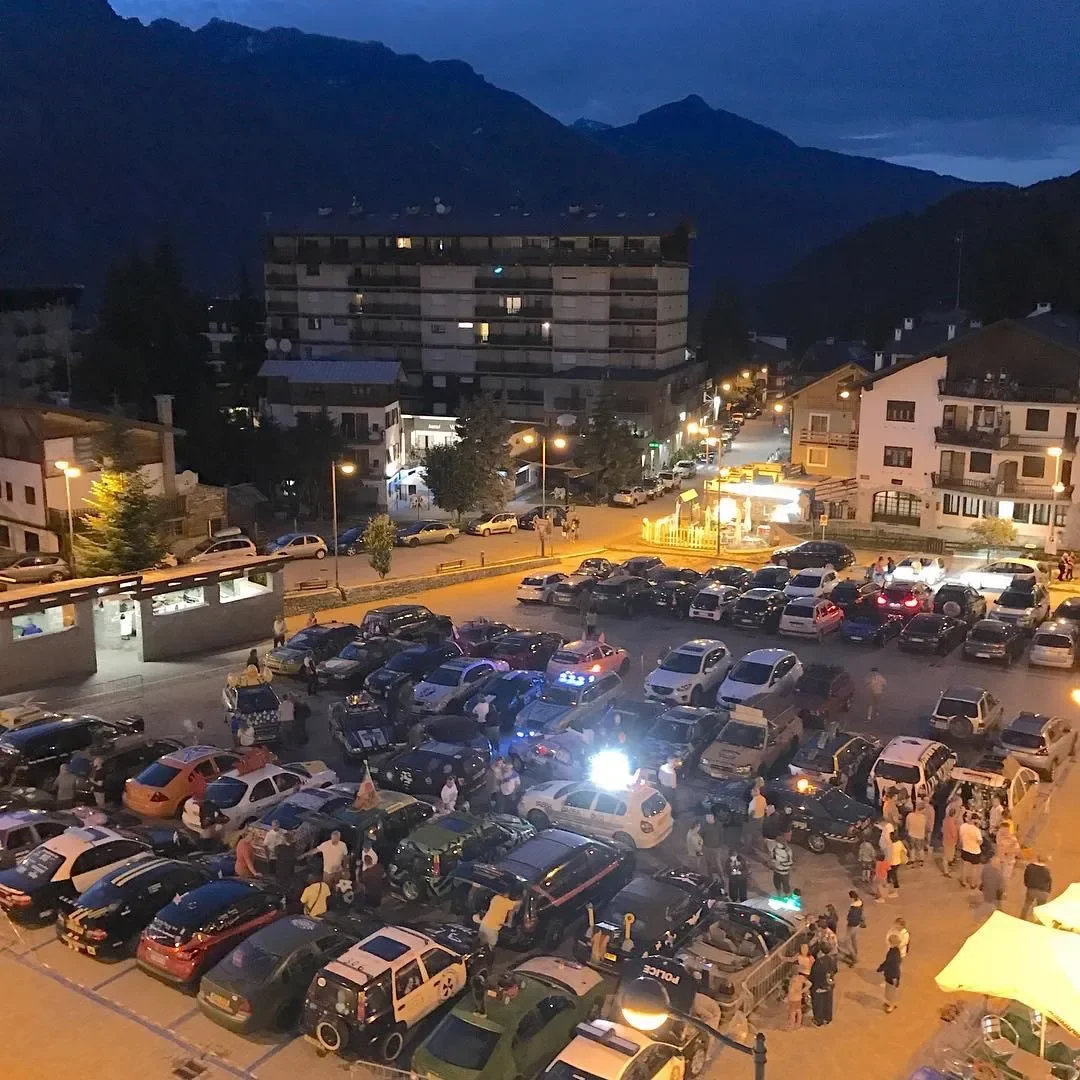 Parking lot filled with cars and people at night, with a mountainous background and buildings illuminated by streetlights.