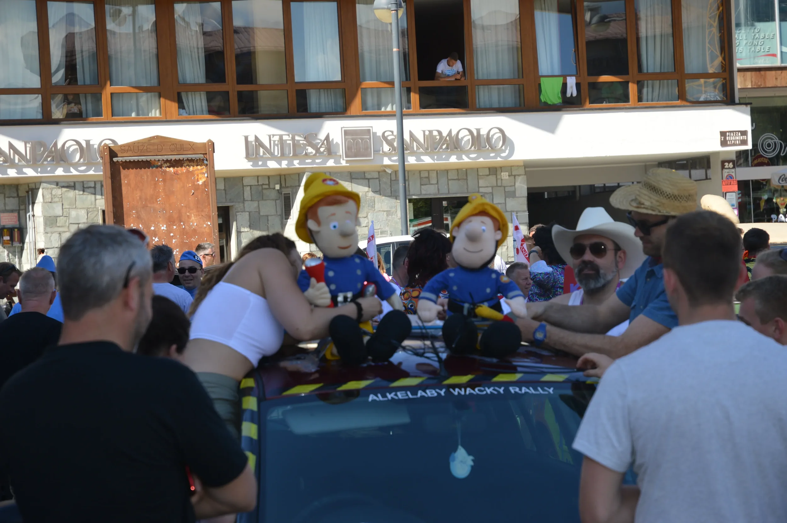 People gather at a rally with large cartoon character puppets on a vehicle, with the text 'Alkelaby Wacky Rally' on the vehicle's windshield. The scene takes place in an urban area, with some participants wearing hats and sunglasses.