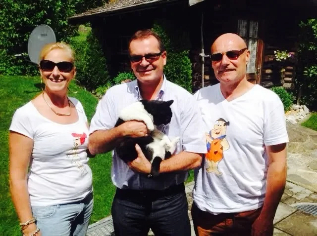 Three adults standing outdoors on a sunny day, smiling at the camera. The person in the middle is holding a black and white puppy. There is a house or shed in the background, surrounded by green grass and some bushes.