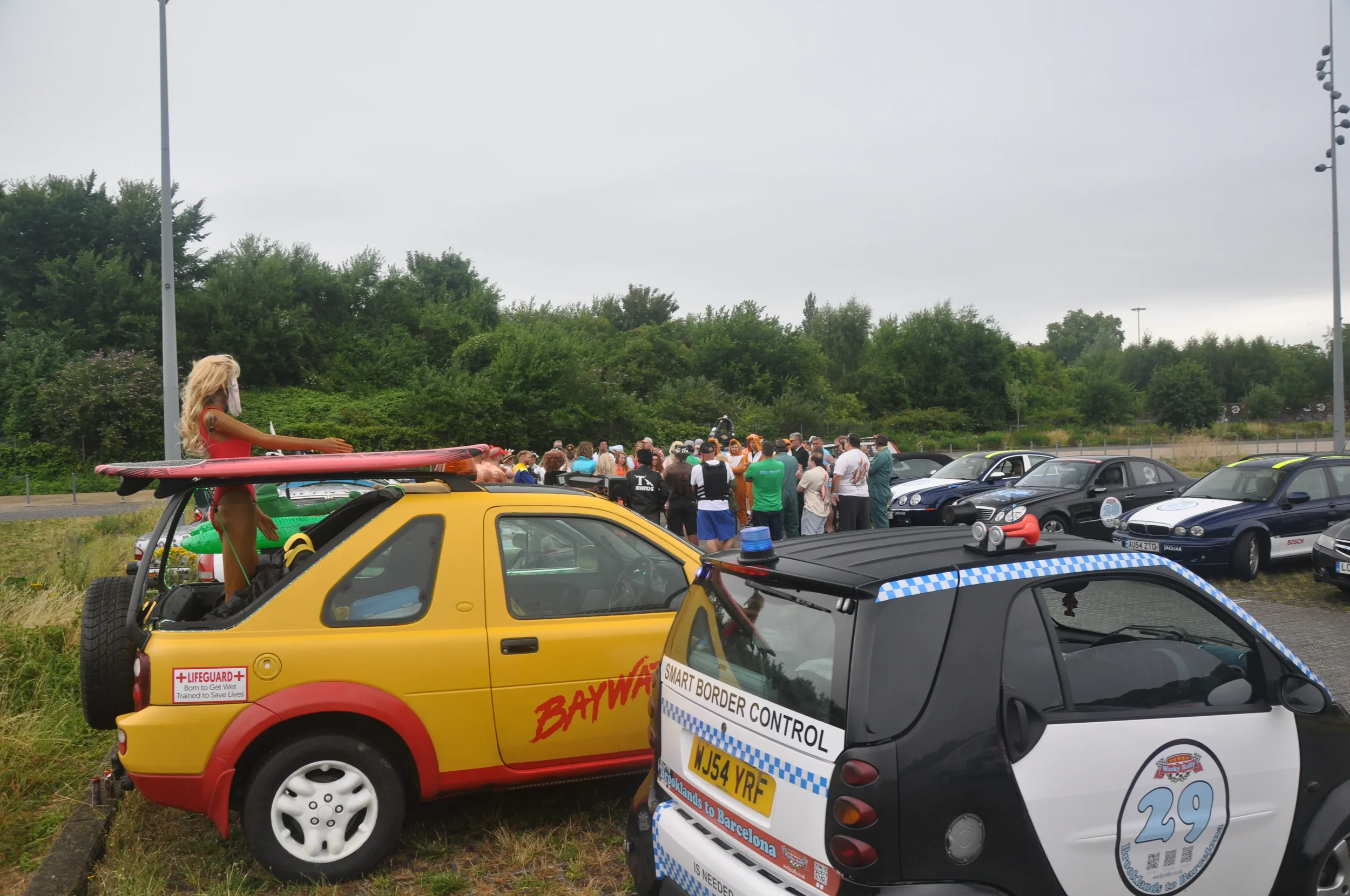 A parking lot filled with various cars, including a yellow vehicle with a surfing dummy on top, and a smaller black car with police markings. People are gathered in the background, some wearing casual clothing and hats, near the edge of the lot.