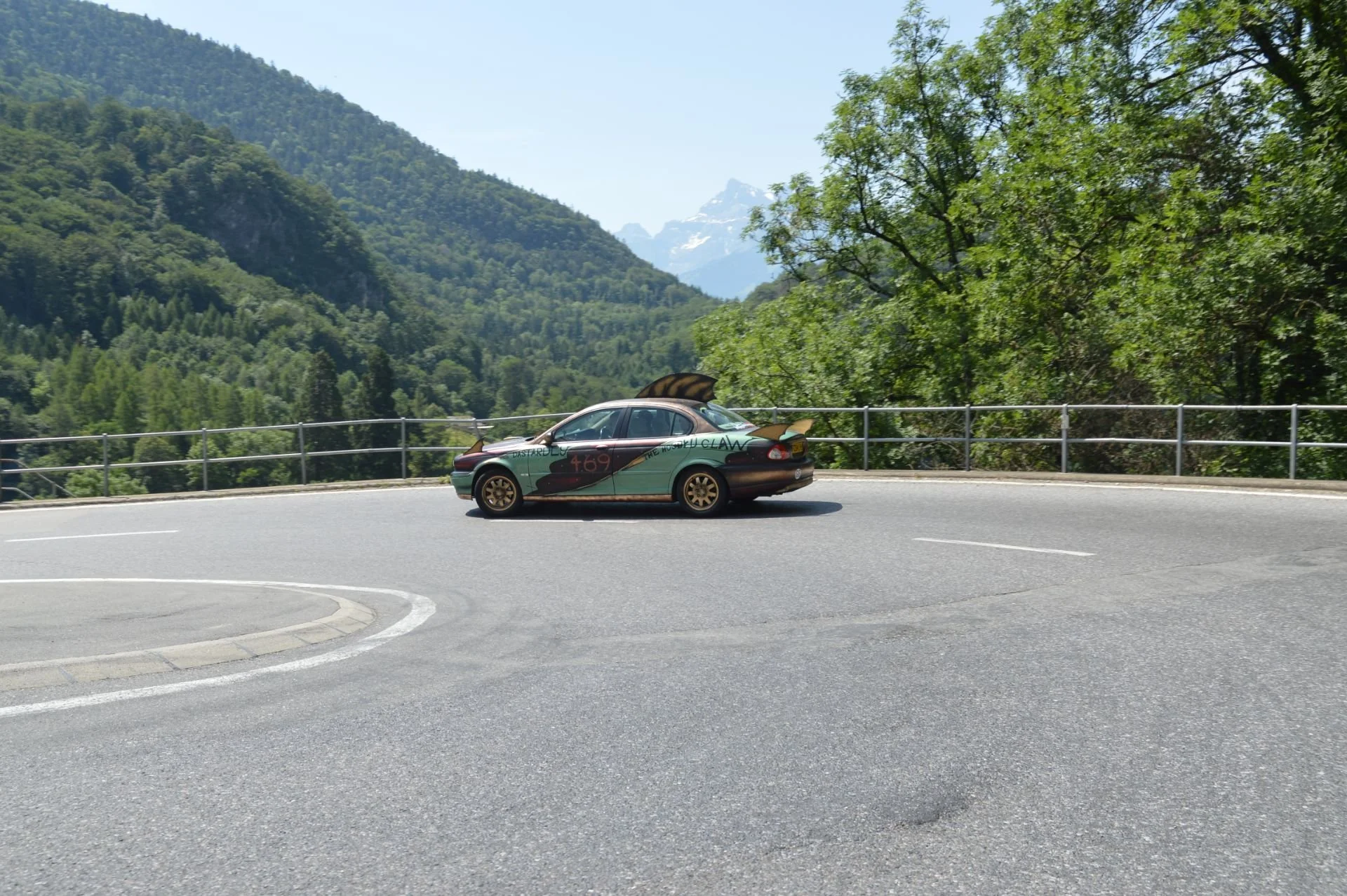 A shiny sports car with painted designs and the number 469 parked on the side of a mountain road surrounded by green trees and distant snow-capped peaks.