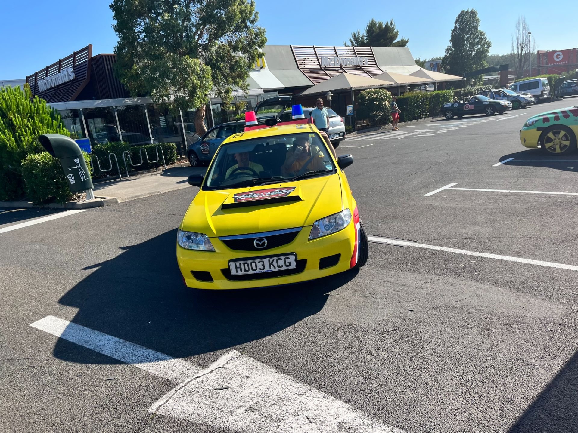 A small yellow car parked in a parking lot outside a McDonald's restaurant, with a woman in the driver's seat and a man next to the car. The car has pink and blue police lights on top.