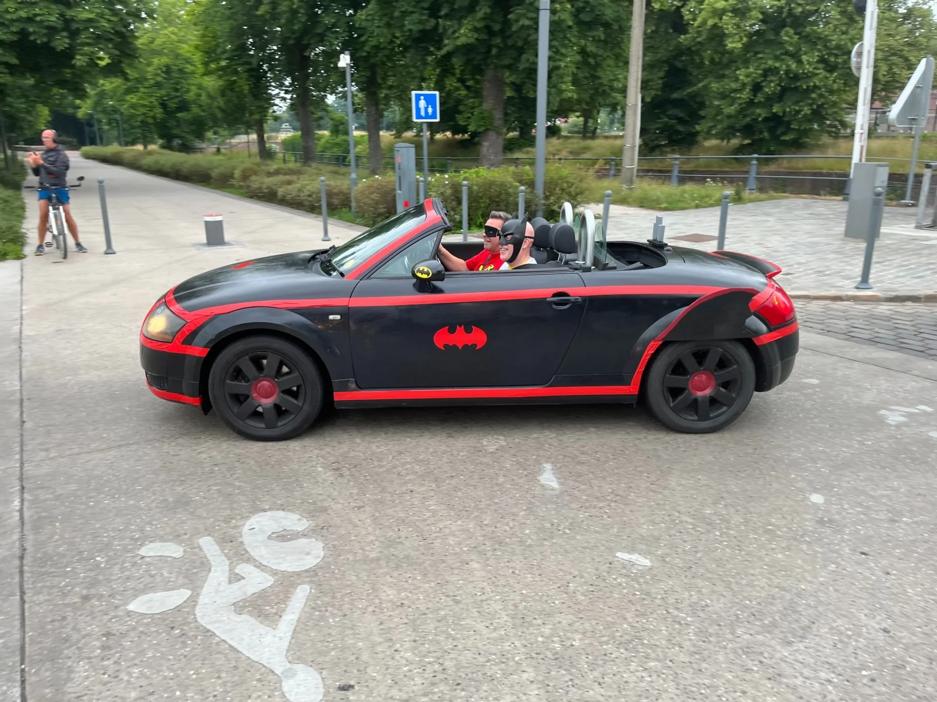 A miniature Batmobile-style car with Batman-themed stickers and two people dressed as Batman and Robin inside, parked on a bike lane near a park with green trees and a person on a bicycle.