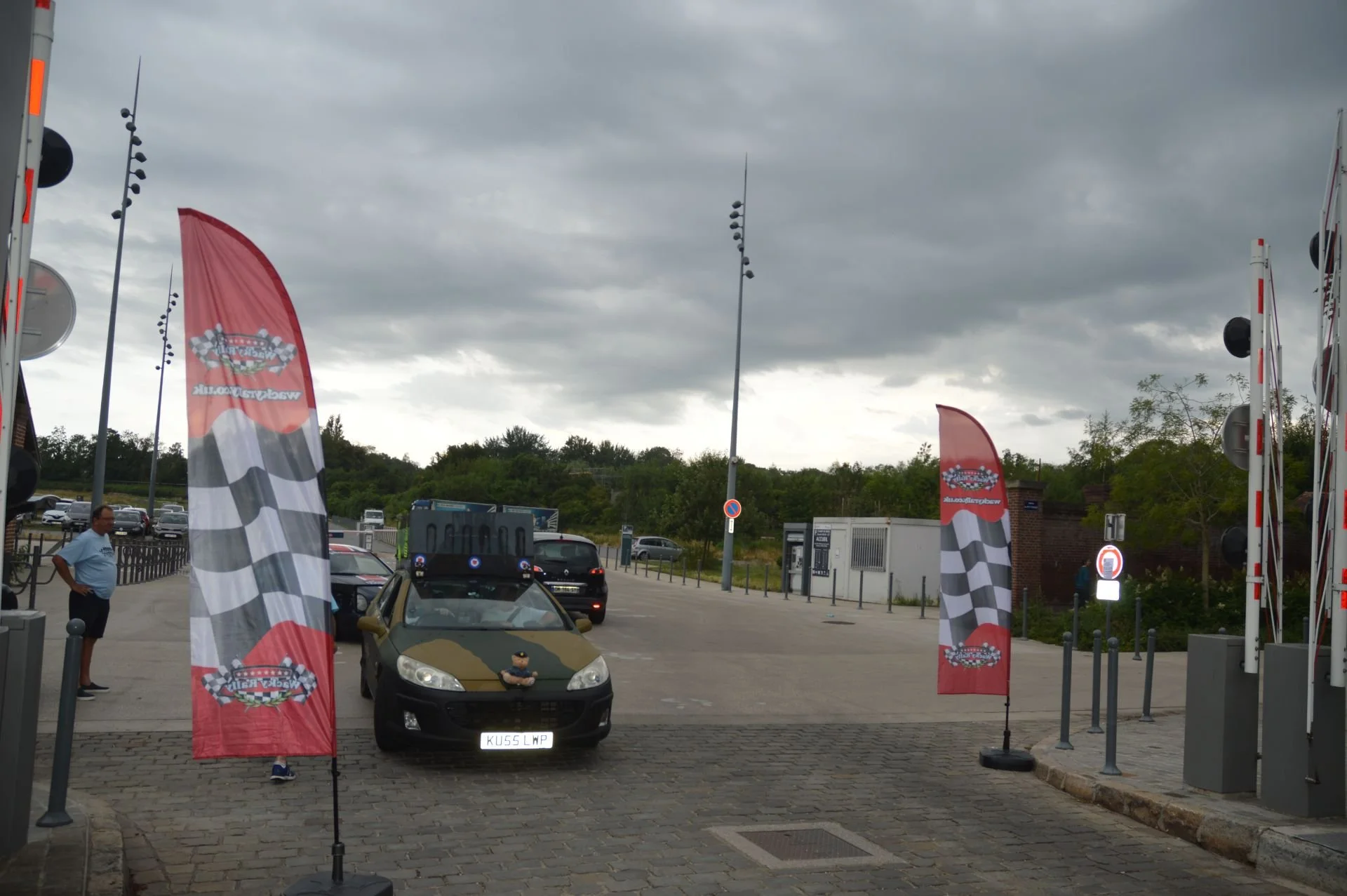 Cars at a racing-themed parking lot with flags and cloudy sky.