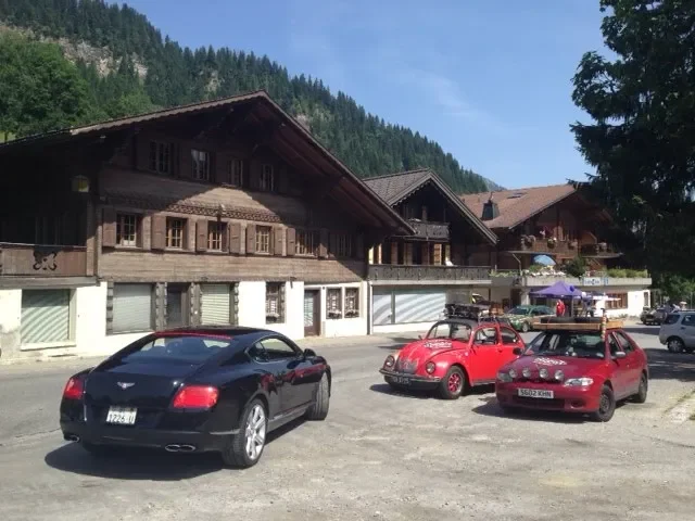 Several parked cars, including a black sports car, a red vintage beetle, and a red hatchback, in front of wooden buildings with mountains and trees in the background.