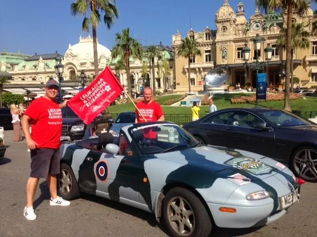 Two men wearing red T-shirts standing next to a camouflage-patterned sports car with a Royal Air Force roundel, holding a red flag that reads 'Lifeline Oklahoma' with a building and palm trees in the background.