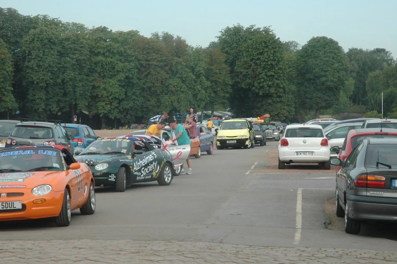Parking lot filled with various cars, some with banners and flags, with people gathered around them, and large green trees in the background.