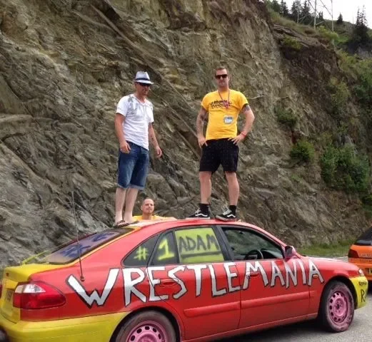 Three men standing on top of a red car with yellow accents, decorated with the words "WrestleMania" and "#1 Adam" on the side, parked outdoors near a rocky hillside.