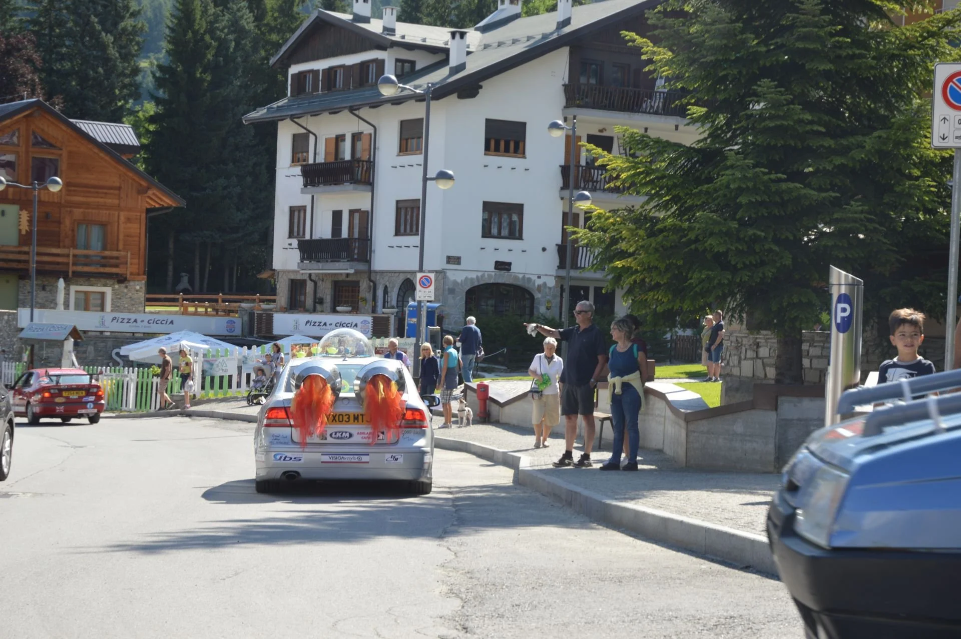 A street scene in a mountain town with people walking and standing along the sidewalk. There is a white car with decorations on the back, possibly for a celebration or parade. The background features a large white building with multiple balconies and