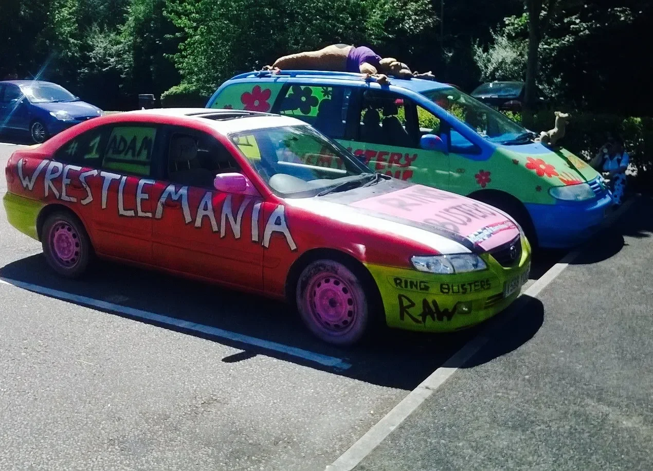 Colorful car with painted words "WrestleMania" on side, parked in a parking lot alongside other decorated vehicles, including one with flower designs and a large stuffed animal on top.