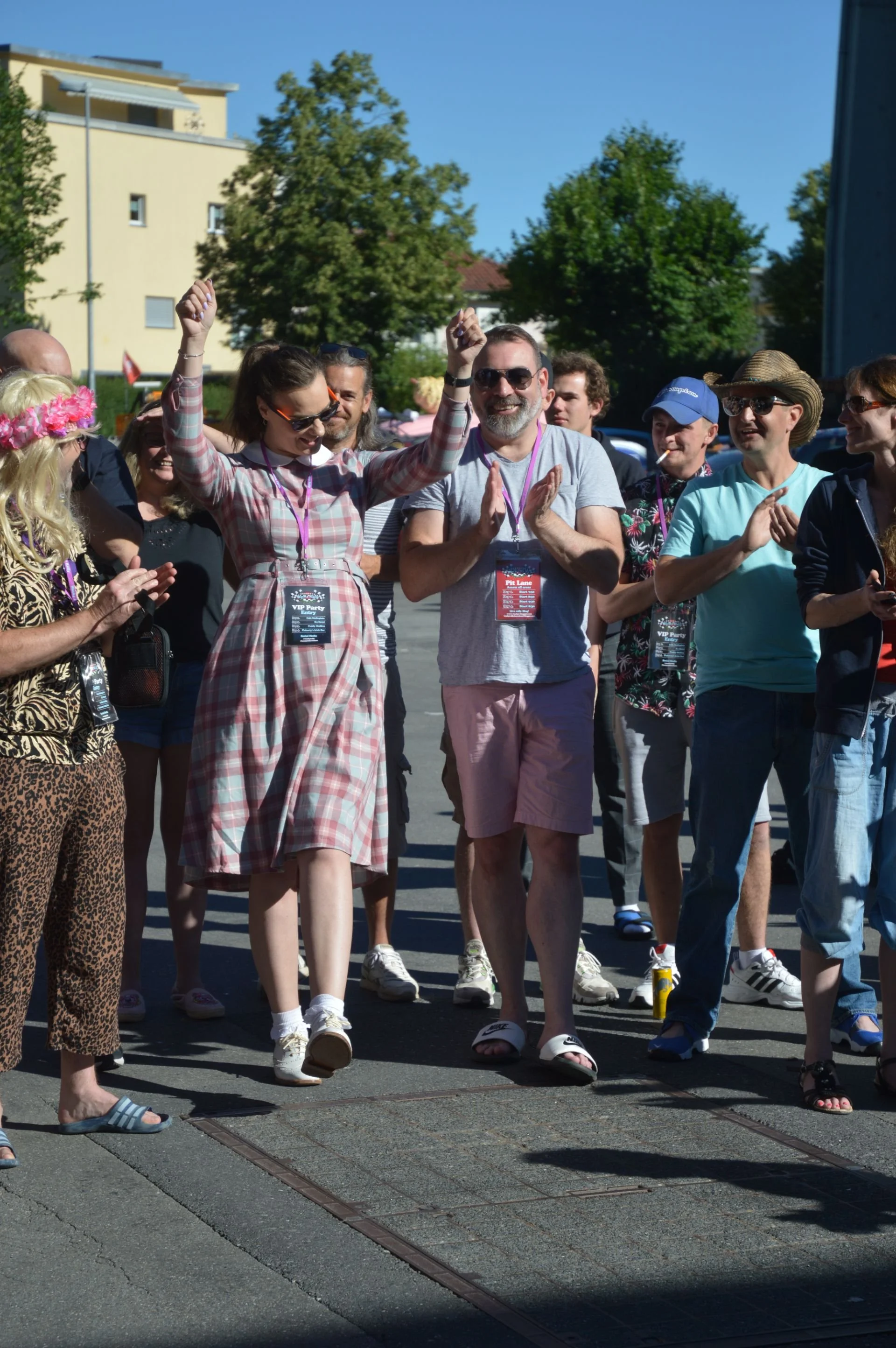 Group of people outdoors on a sunny day, celebrating or enjoying an event. A woman in a plaid dress is dancing or jumping, while others are clapping and smiling.