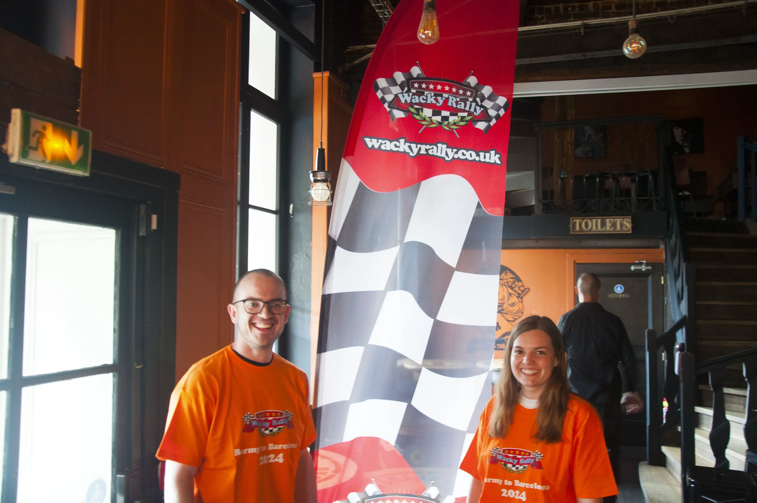 Two smiling people wearing orange t-shirts, standing indoors near a red, black, and white checkered flag and a banner that reads 'Wacky Rally' and 'wackyrally.co.uk'. The background features a staircase, a 'TOILETS' sign, and an exit door.