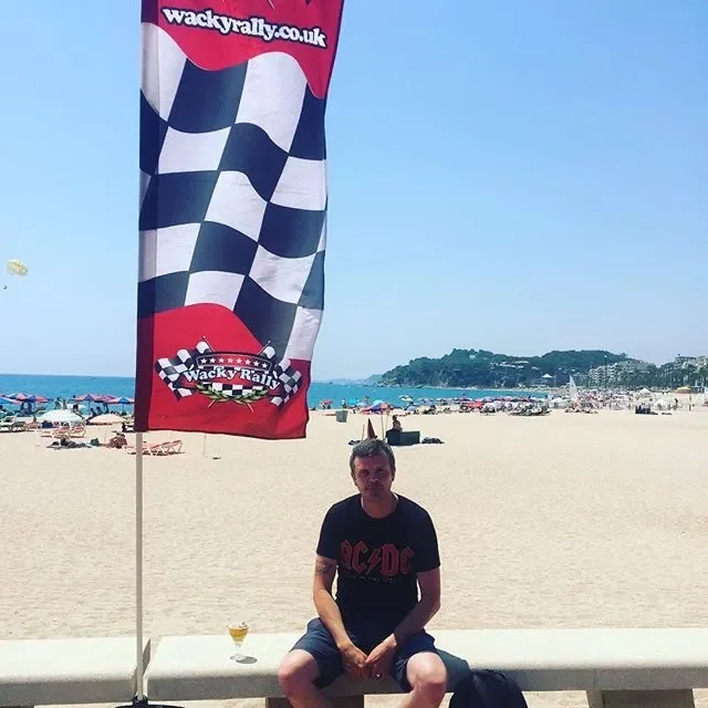Man sitting on a concrete bench at the beach near a wacky rally flag with a checkered pattern and the website "wackyrally.co.uk" printed on it.