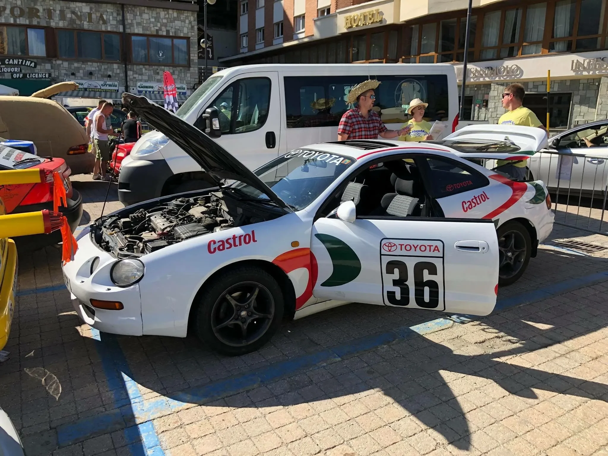 White Toyota race car with the hood open, displaying its engine. The car has the number 36 and is sponsored by Castrol. It is parked on a paved area next to other cars, with people and buildings in the background.
