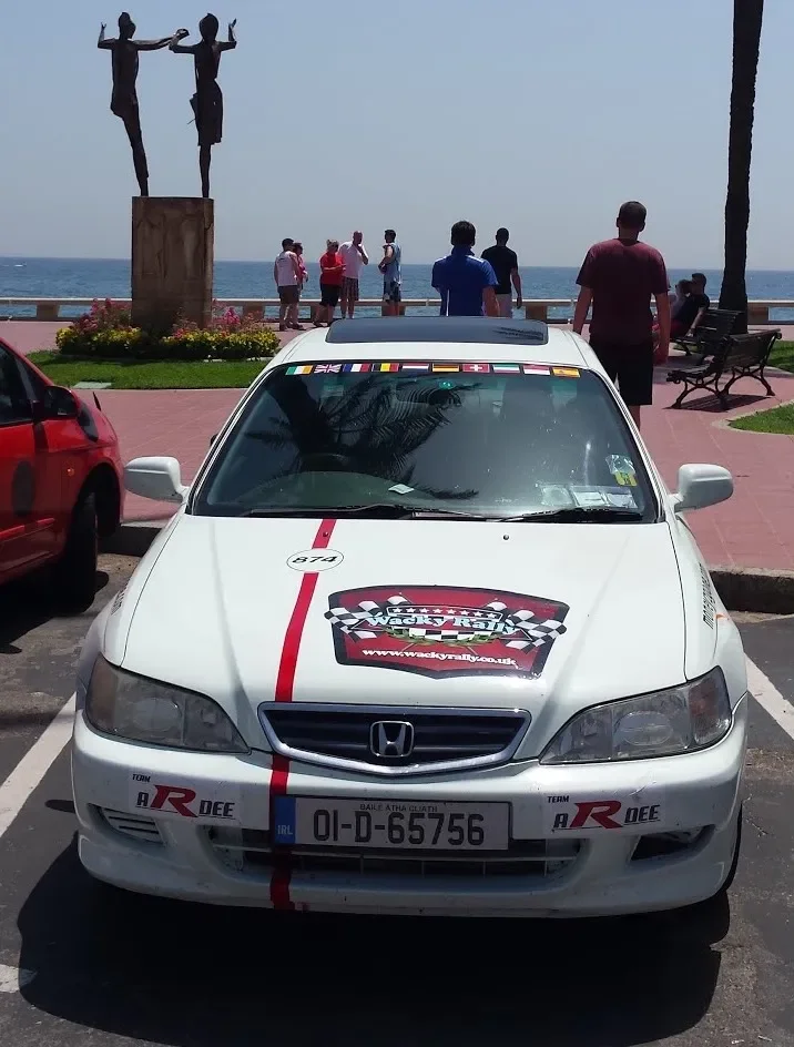 A white Honda car with racing decals parked in a lot near a seaside promenade. The front of the car displays the website 'www.wackyrail.co.uk' and logos indicating participation in rally racing. There are several people in the background looking at t