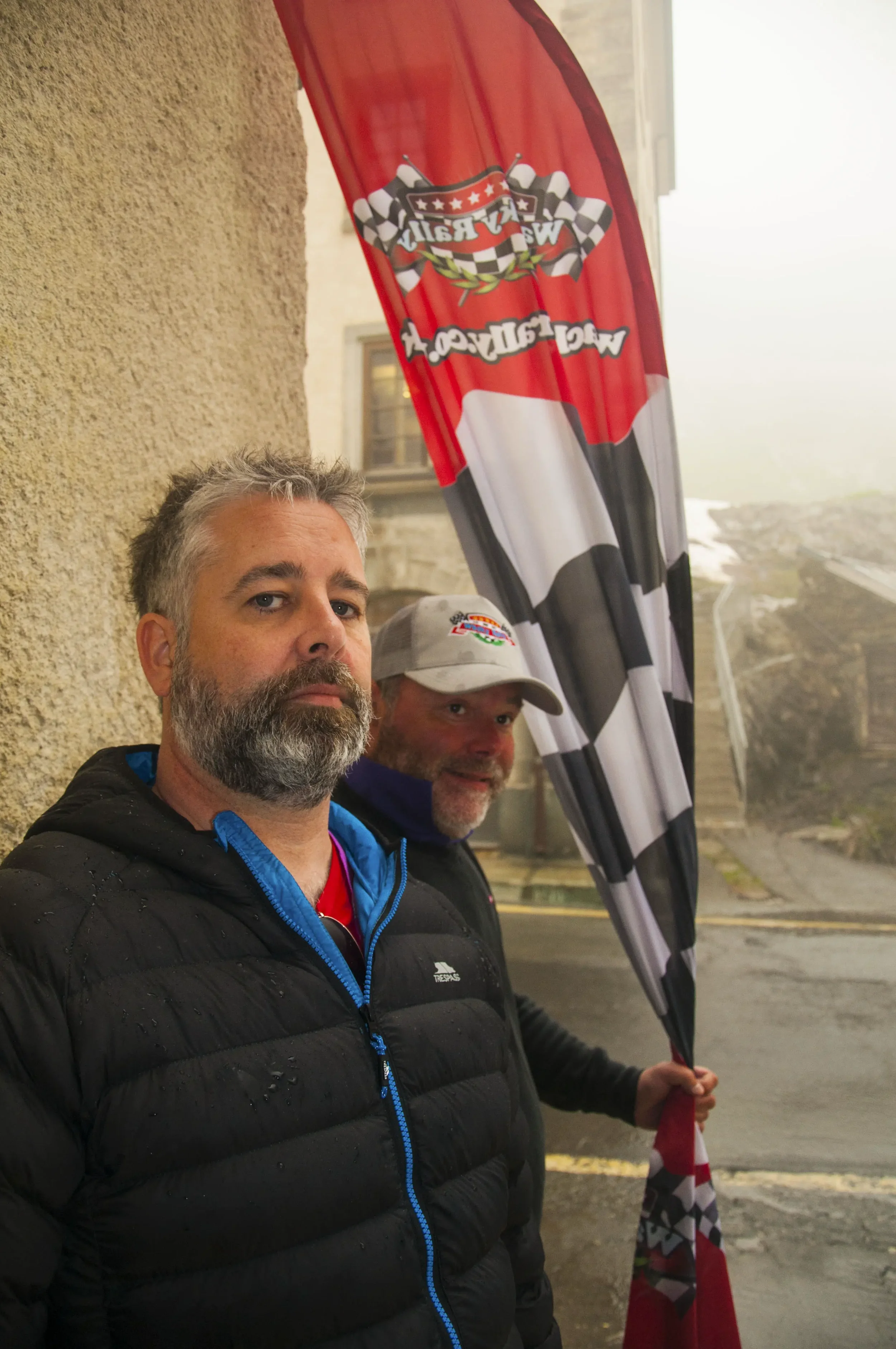 Two men standing outside near a building under a waving checkered flag and a vertical red and black banner with racing-related graphics.