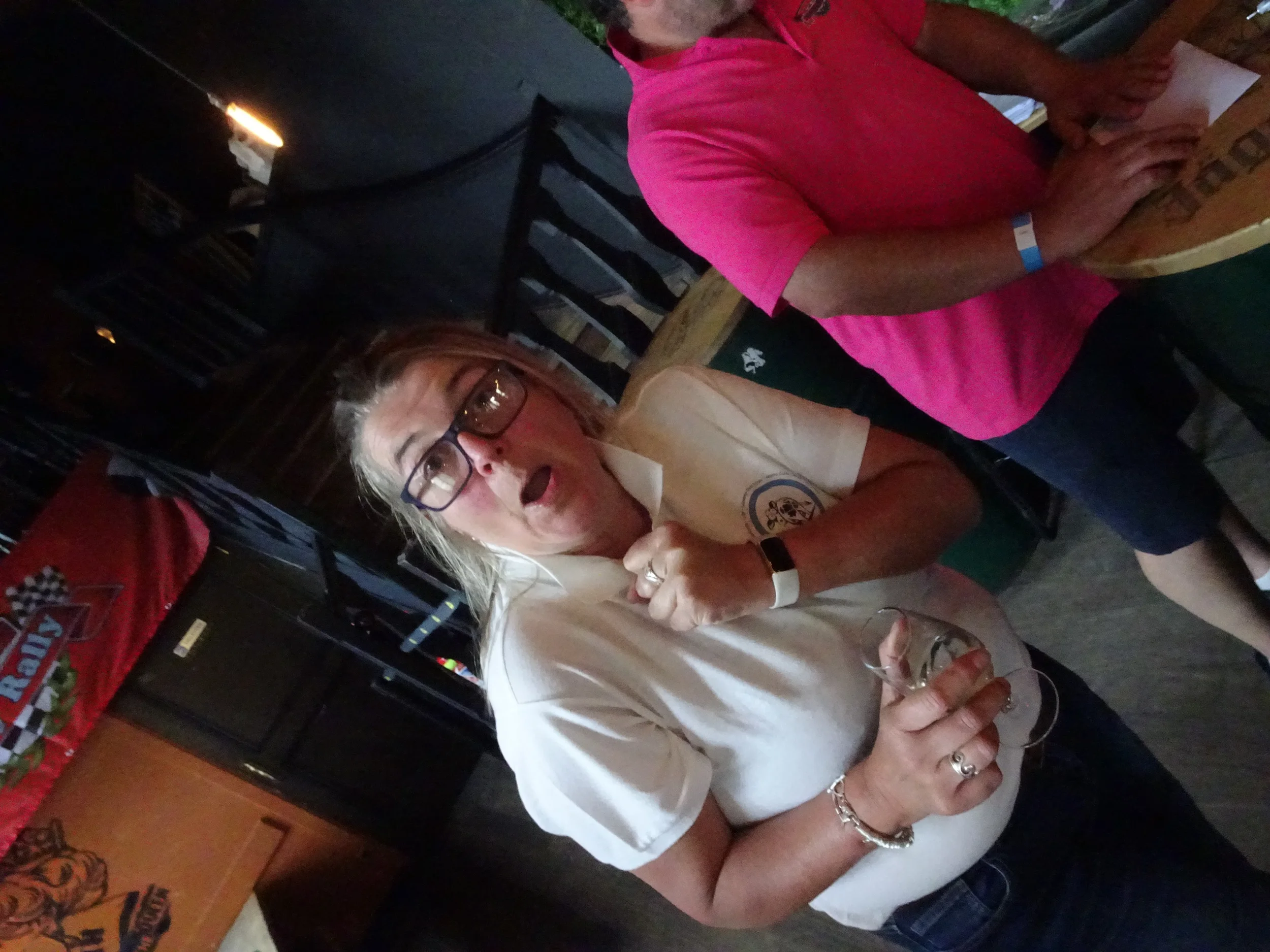 A woman with glasses and blonde hair making a surprised or playful expression while holding a glass of wine. She is wearing a white shirt and standing indoors at what appears to be a social gathering or event.