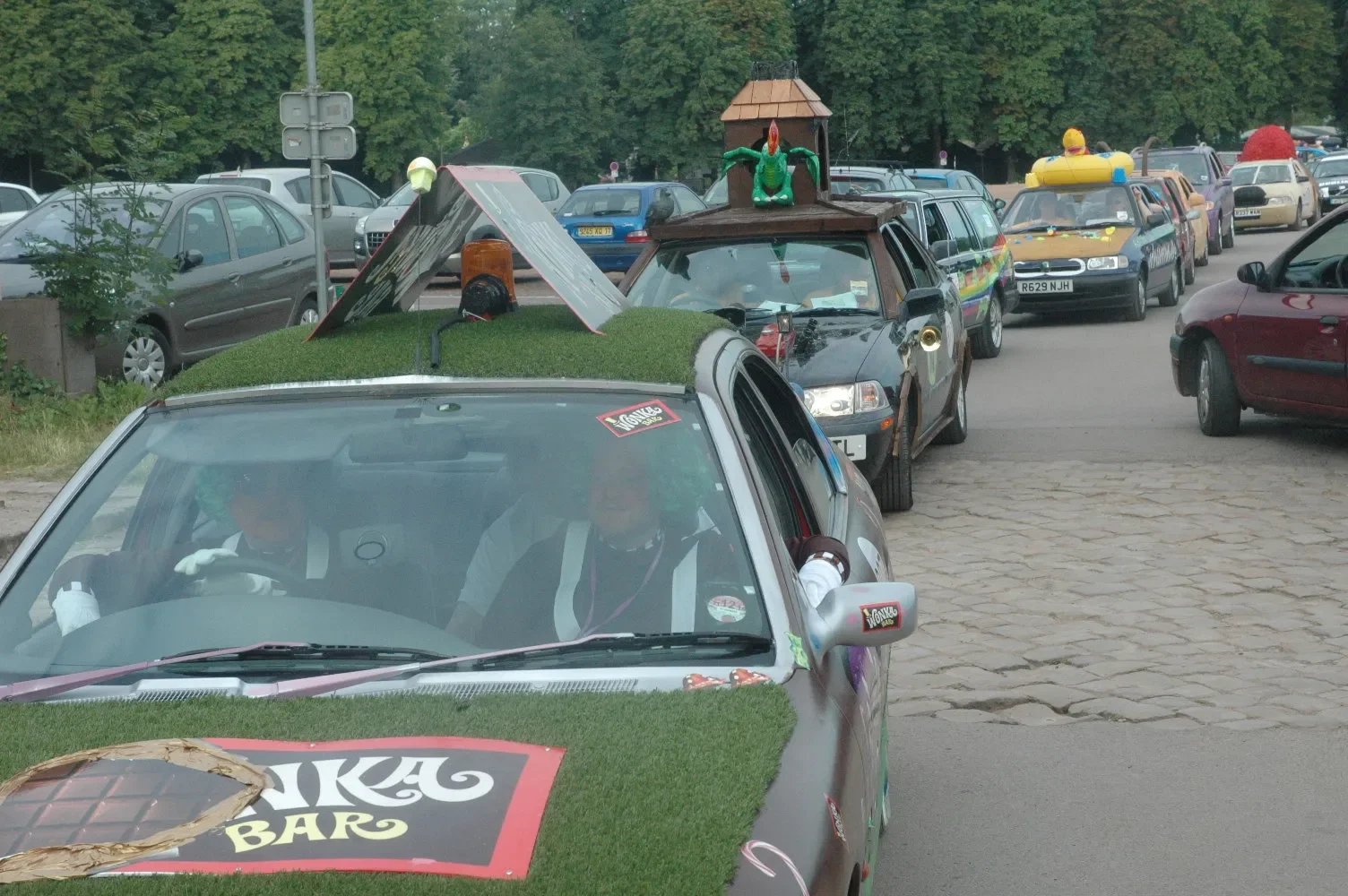 Line of decorated cars with various props and decorations, likely for a parade or event, on a paved street with parked cars and trees in the background.