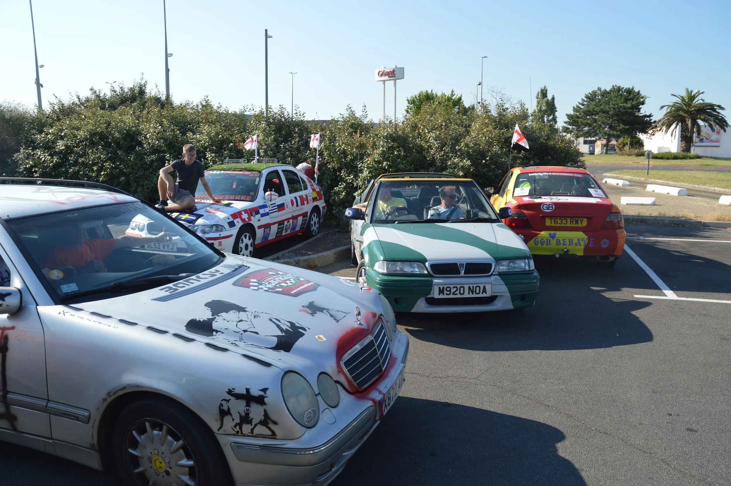 Cars decorated with flags and stickers parked in a lot, with people sitting inside and outside, under a clear sky.