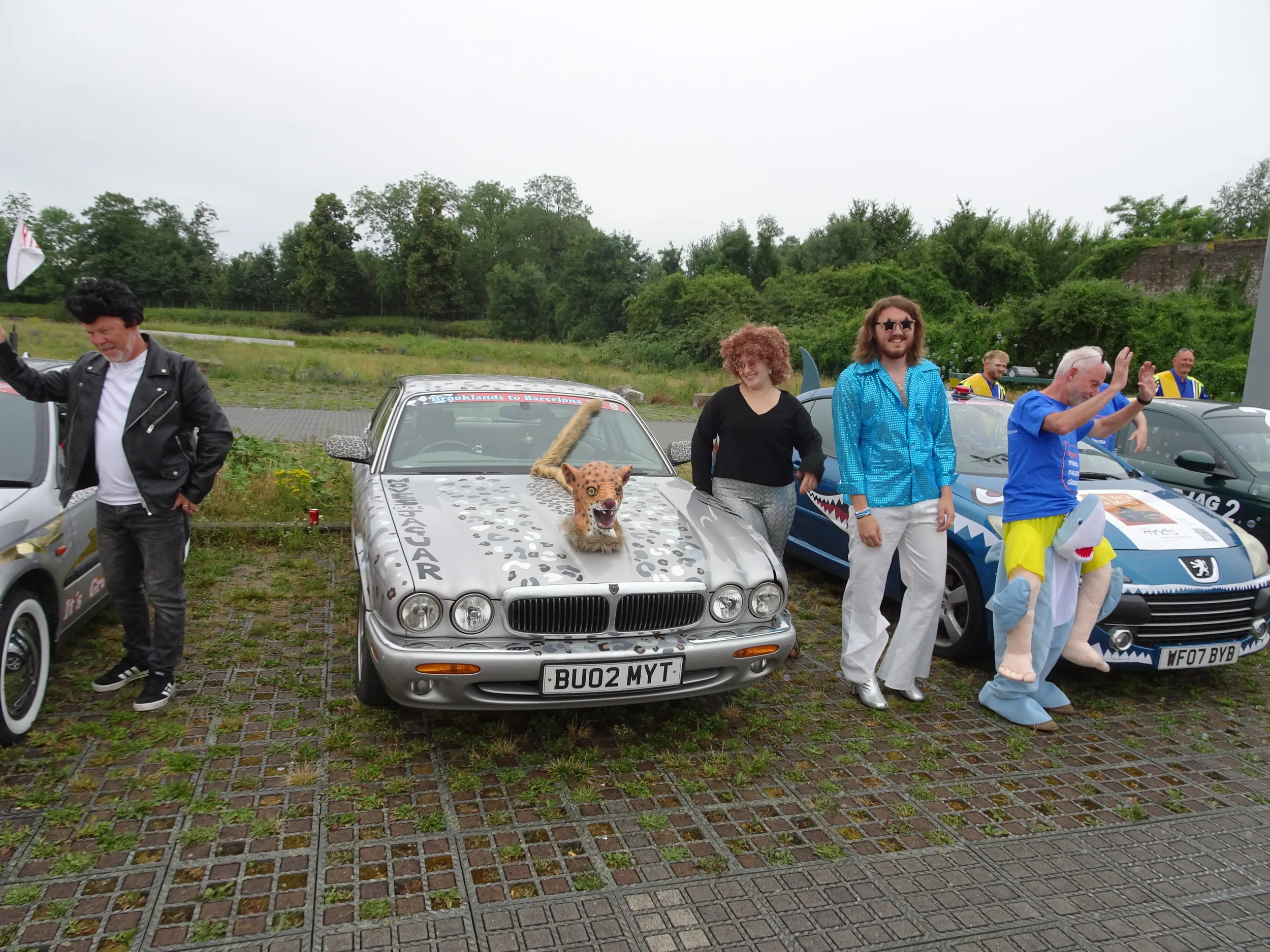 People standing next to parked cars at a car event, with one car decorated to resemble a jaguar and another with shark-themed decorations.
