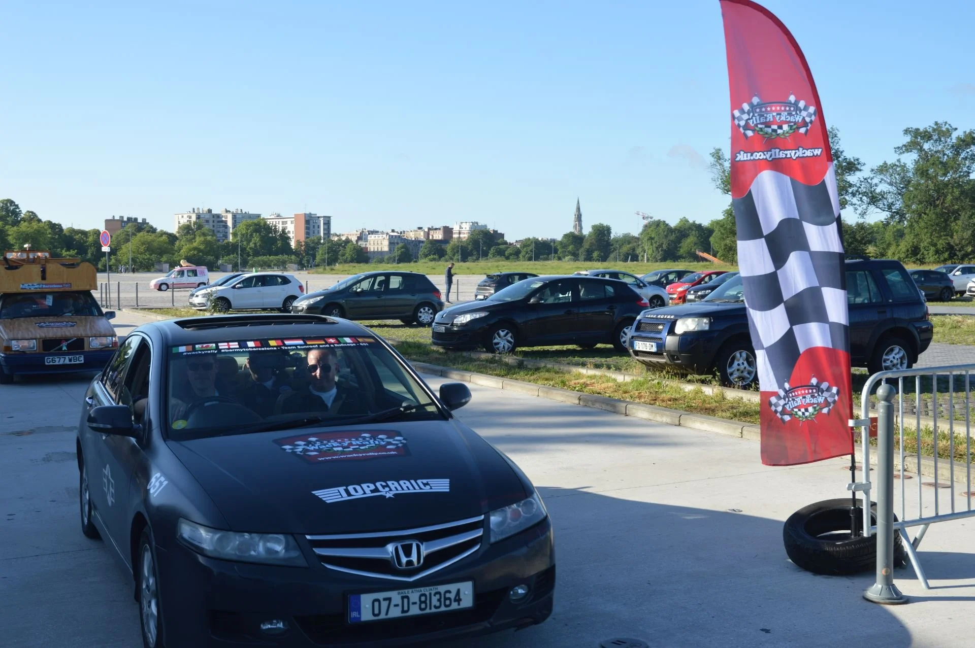 A black Honda car with a racing-themed decal on the hood and windshield, parked near a tall checkered flag banner. The background shows a parking lot with several other cars, a few buildings, and a clear blue sky.