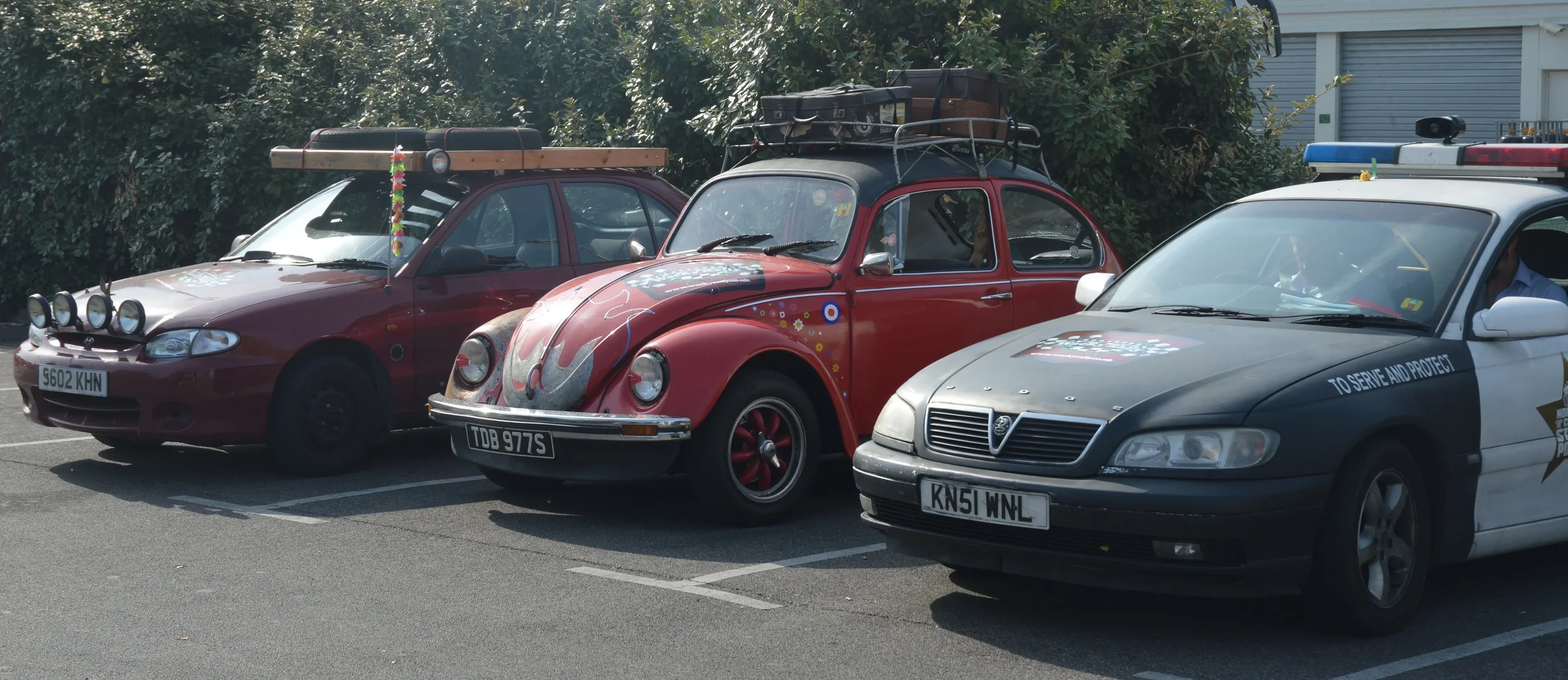 A lineup of three cars parked in a lot, including a red Volkswagen Beetle with roof luggage, a maroon hatchback with off-road lights, and a police car with police markings and emergency lights.