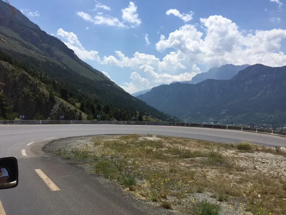Curving mountain road with guardrail, overlooking a valley with mountains and a partly cloudy sky in the background.
