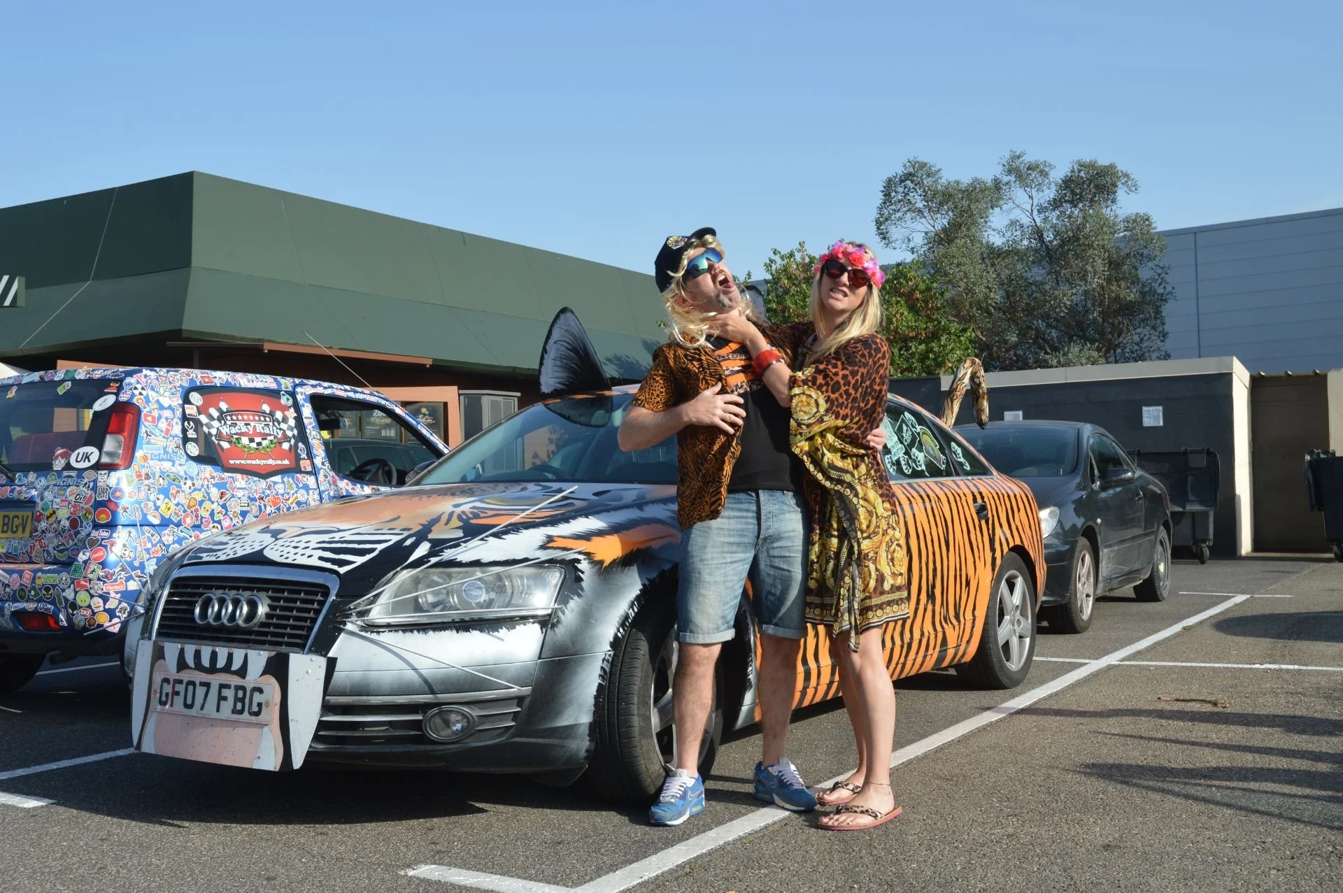 Two people dressed in animal print costumes stand in front of a decorated car in a parking lot, with one wearing a hat and the other a pink floral headband, both wearing sunglasses and striking playful poses.