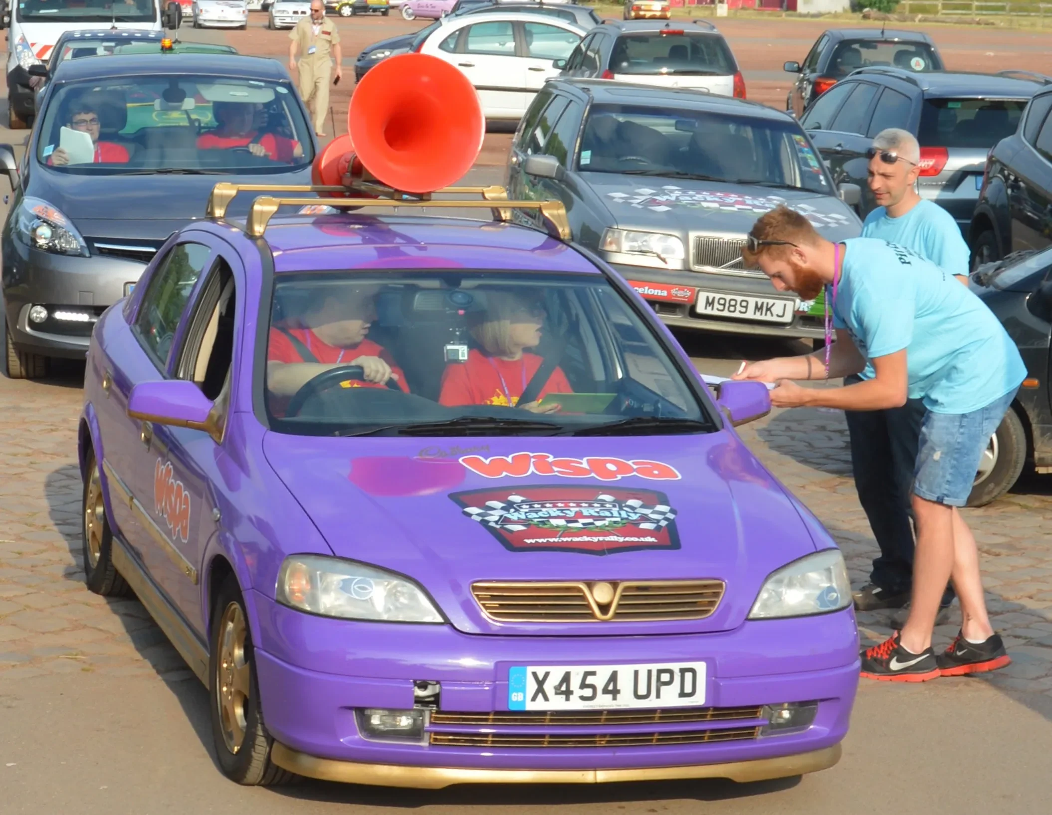 Purple race car with a large red megaphone on its roof, parked in a lot with multiple cars around. Two children are inside the car wearing red shirts, and an adult in a blue shirt is leaning into the driver's side window, talking to them.