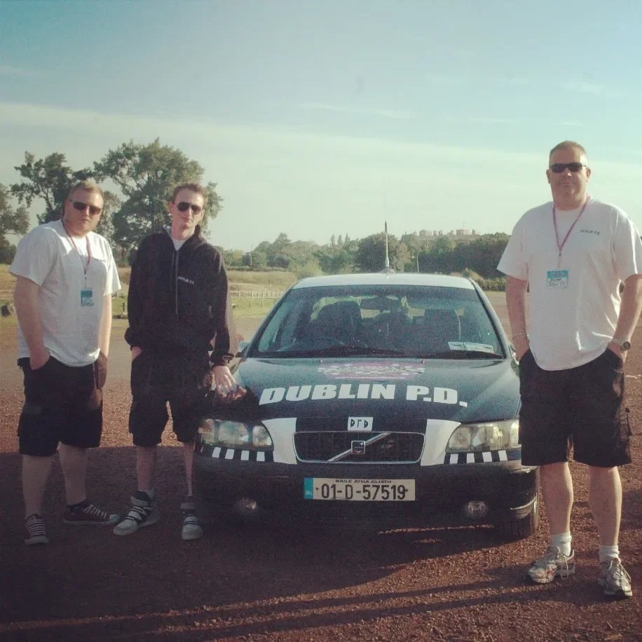 Three people standing next to a black and white Dublin Police car in an open field during daytime, wearing casual clothing and sunglasses.
