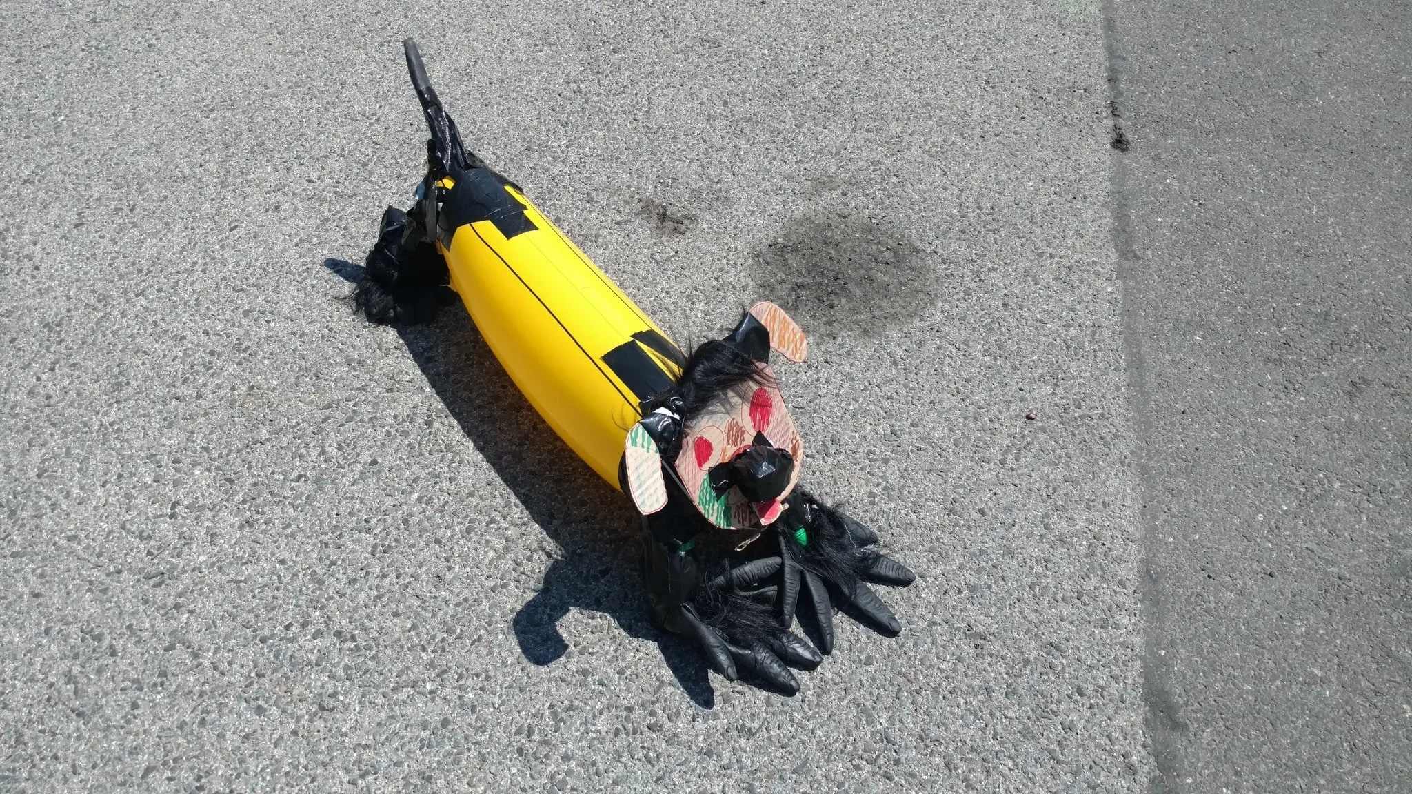 A handcrafted dog sculpture made from a yellow plastic bottle, black gloves, and various craft materials is lying on asphalt pavement.