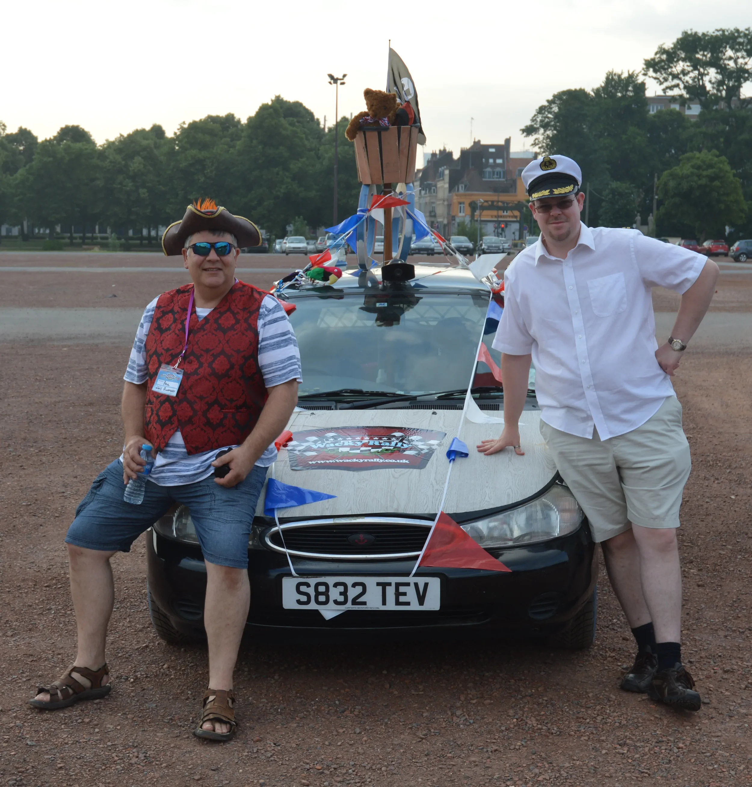 Two men dressed in celebratory costumes posing in front of a decorated car with a teddy bear in a bucket on top, in an open parking lot during daytime.