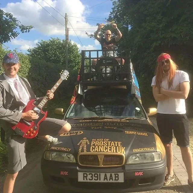 Three people posing with a decorated black car; a man on the left in casual gray clothing and a baseball cap holding a guitar, a woman on the right in a white t-shirt and black shorts standing with arms crossed, and a man on top of the car holding a 
