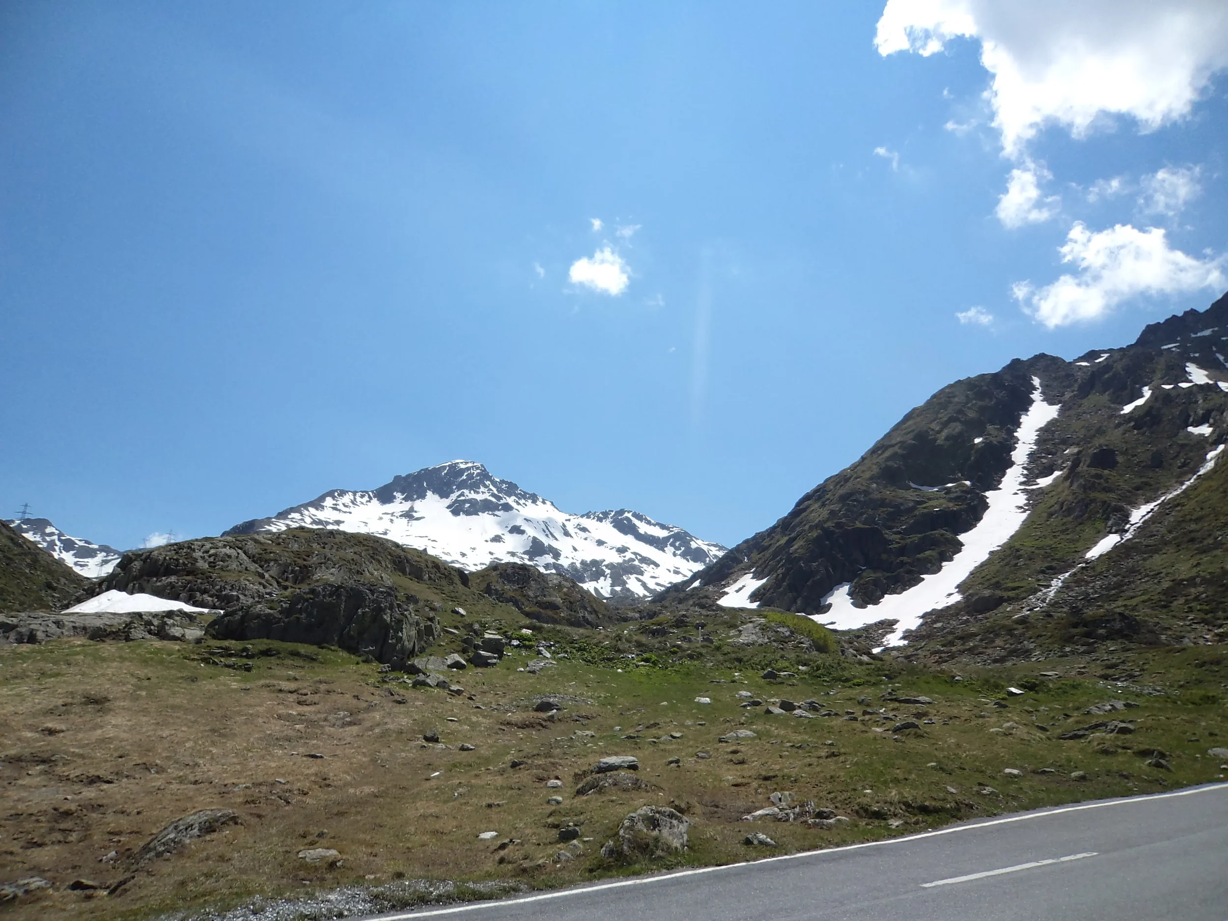 Scenic mountain landscape with snow-capped peaks, grassy slopes, and a road in the foreground under a bright blue sky with a few clouds.